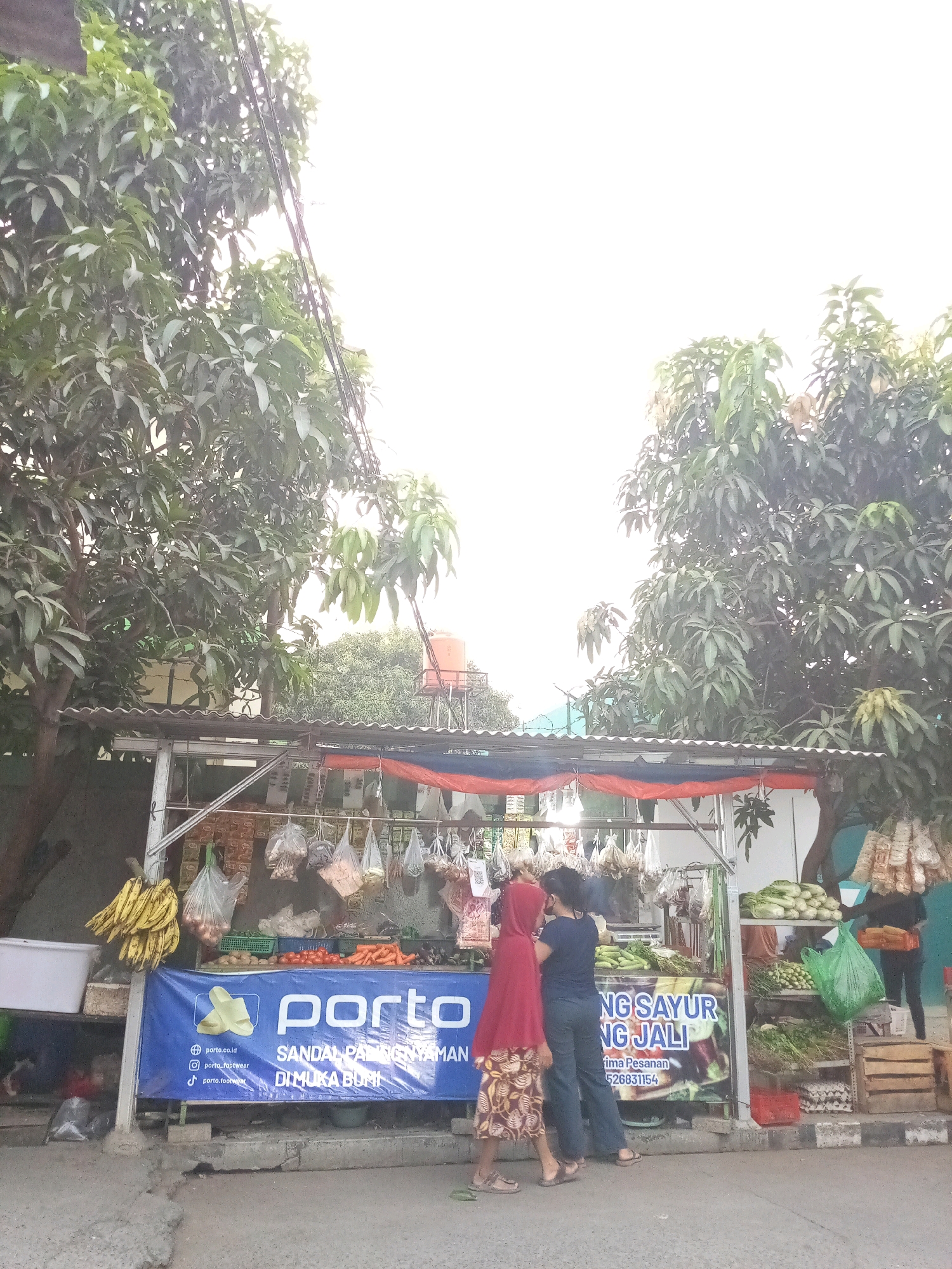 This is a street-level, eye-level, medium shot image of an outdoor market stall in Bekasi, Indonesia. The stall is set up under a corrugated metal roof supported by metal poles. The roof is adorned with red and blue tarps. The stall is laden with various fresh produce including bananas, onions, tomatoes, carrots, and leafy greens. Bags of what appear to be dried goods or spices are hanging from the stall. A large blue banner with the "porto" logo and text in Indonesian, advertising "SANDAL PADANYA NYAMAN DI MUKA BUMI" and a phone number, is displayed prominently in front of the stall.
Two individuals are at the stall, appearing to be customers. One, wearing a red headscarf and patterned skirt, stands with their back to the camera. The second individual, in a dark blue top and dark pants, is facing the first person, suggesting an interaction. To the right of the stall, another person in dark clothing is visible, holding an orange crate filled with produce. Wooden crates and egg cartons are stacked nearby. The background shows foliage from trees, power lines, and a water tank on a platform, typical of urban Indonesian scenery. The ground is asphalt, with a concrete curb and a drainage ditch. The sky is bright, suggesting daytime.