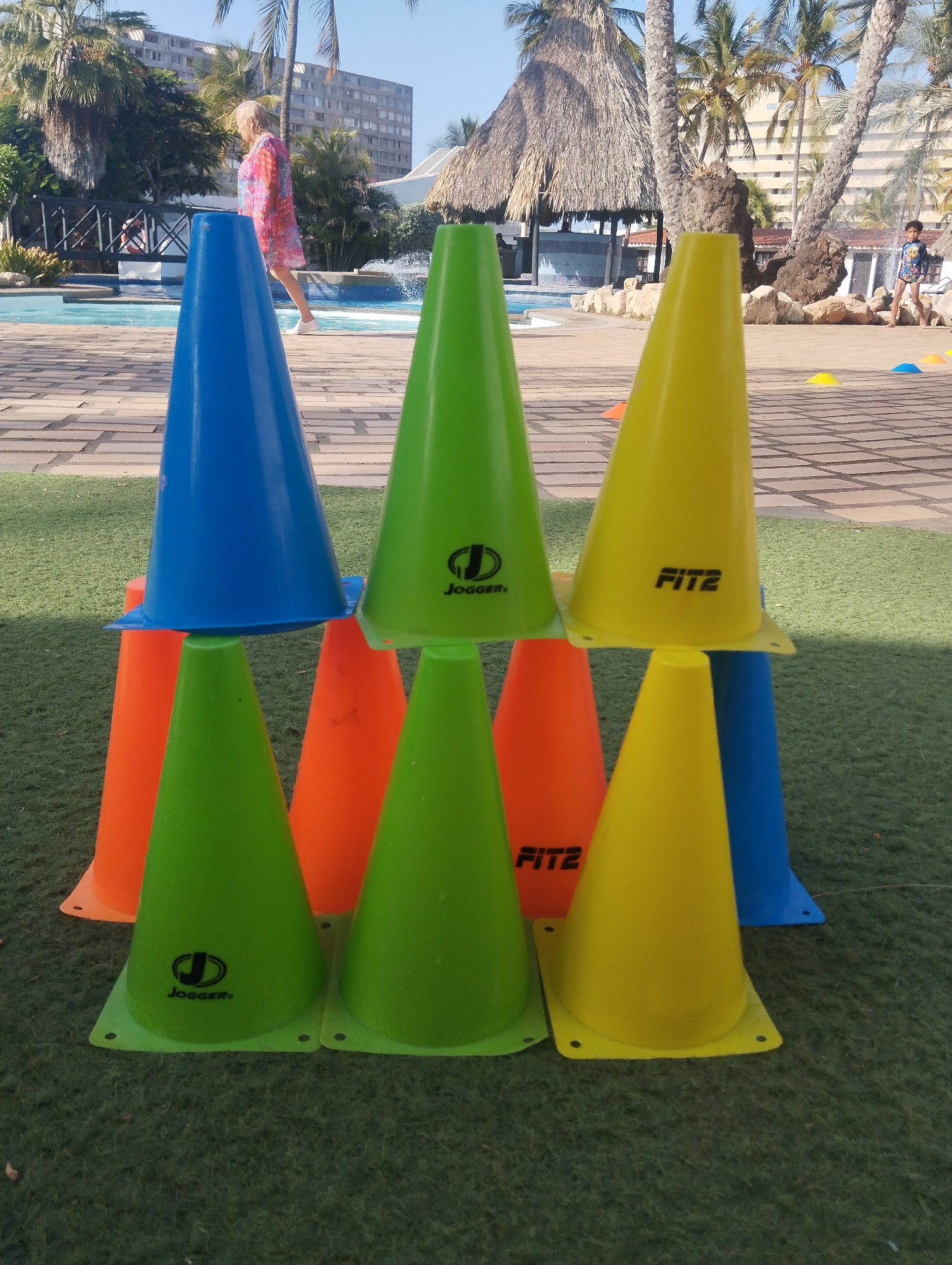 The image captures a stack of traffic cones, arranged in a pyramid formation, on a grassy surface. The cones are in blue, green, yellow, and orange colors, bearing logos "Jogger" and "FITZ" on some. In the background, there is a tropical resort setting with palm trees, a thatched-roof structure, a swimming pool, and a multi-story building. A woman in a pink dress is walking in the background near the pool and bridge. A boy is standing in the distance to the right. The scene appears to be in a tourist area. This image was taken in Porlamar, Venezuela.