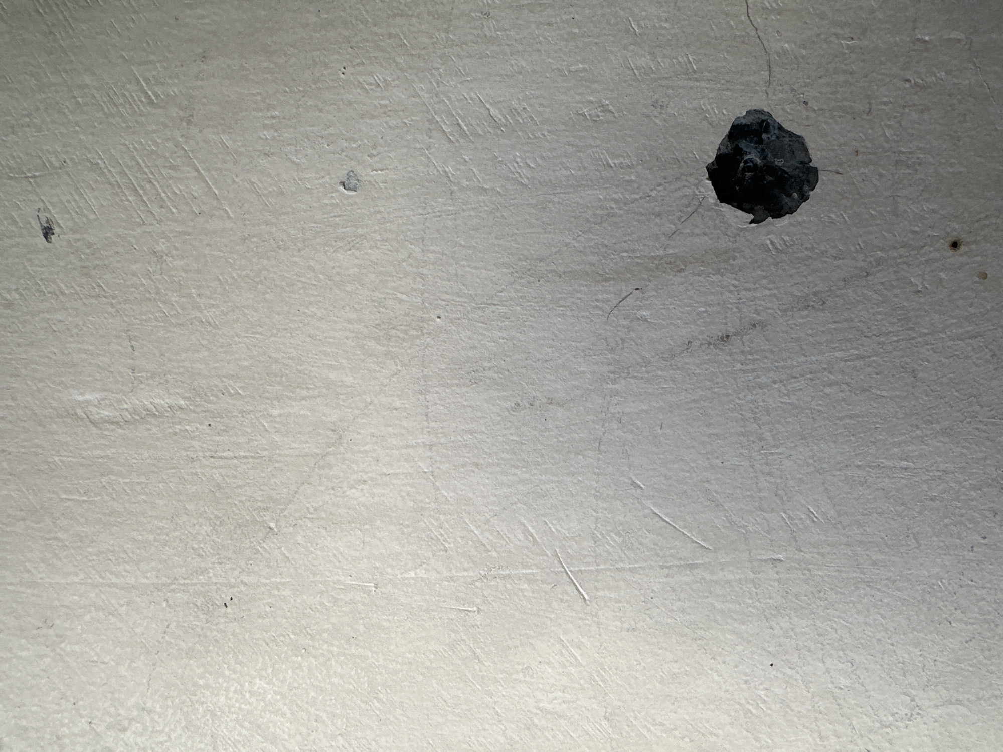 The image shows a close-up, slightly angled view of a textured, off-white wall. The wall surface is not smooth and exhibits numerous fine scratches and some subtle lines. There are a few small dark spots scattered across the wall, likely dust or minor blemishes. In the lower right quadrant, there is a distinct hole or gouge in the wall, which appears dark and irregular, revealing a deeper, shadowed cavity. The lighting suggests an indoor environment, possibly with a single light source casting subtle shadows that accentuate the wall's texture and the hole. No people or specific city landmarks are visible. The context provided about Bulakrejo, Indonesia, does not appear to be directly discernible from the visual information in the image itself, which focuses solely on the texture and damage of a wall.