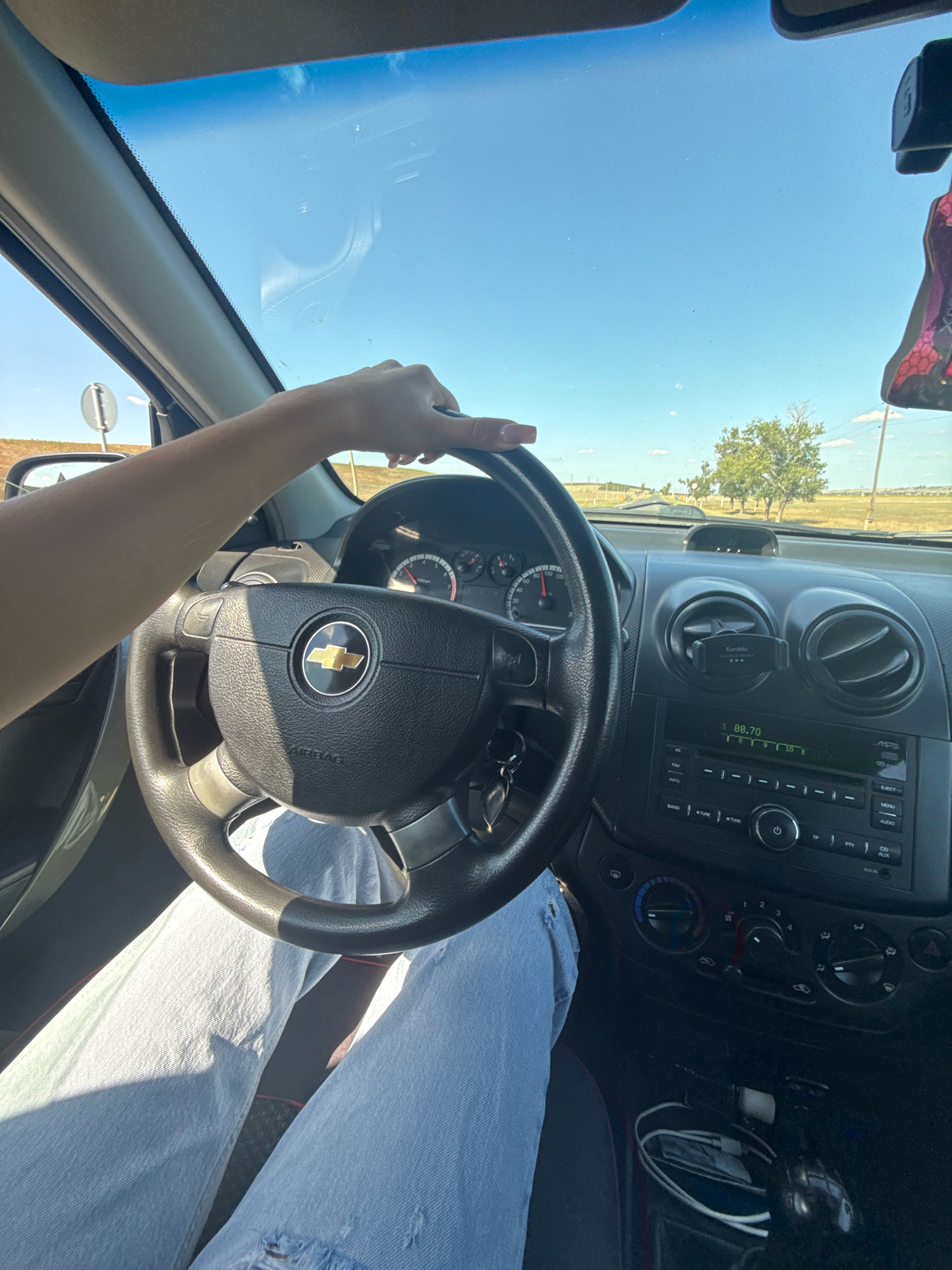 The image shows a person's hands on the steering wheel of a car, suggesting they are driving. The interior of the car is visible, including the dashboard with the radio and air vents. The exterior view from the windshield shows a sunny day with a road stretching into the distance and trees on the side. A traffic sign is visible to the left. The person is wearing light-colored jeans. The image provides a first-person perspective of driving in a rural or suburban area.