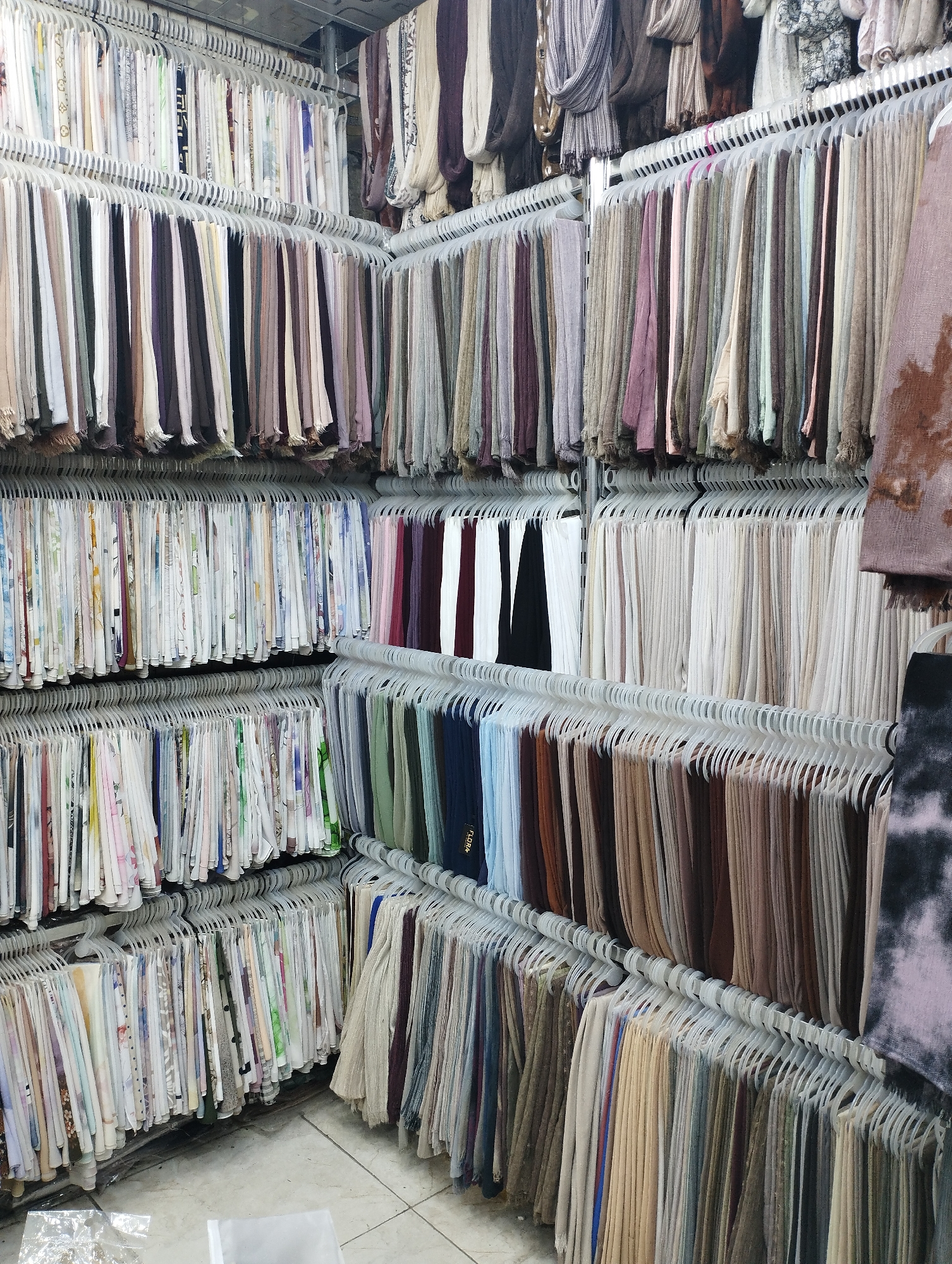 This image depicts a retail store in the city of Baqa'a camp, Jordan, specializing in textiles, likely scarves or fabric. The store is filled with rows of clothing items, predominantly scarves, neatly displayed on white hangers and arranged on multiple tiers of shelving. The items vary in color, pattern, and texture, suggesting a wide selection for customers. The lighting in the store appears to be overhead and functional, illuminating the merchandise. The floor is tiled. No people are visible in the image.