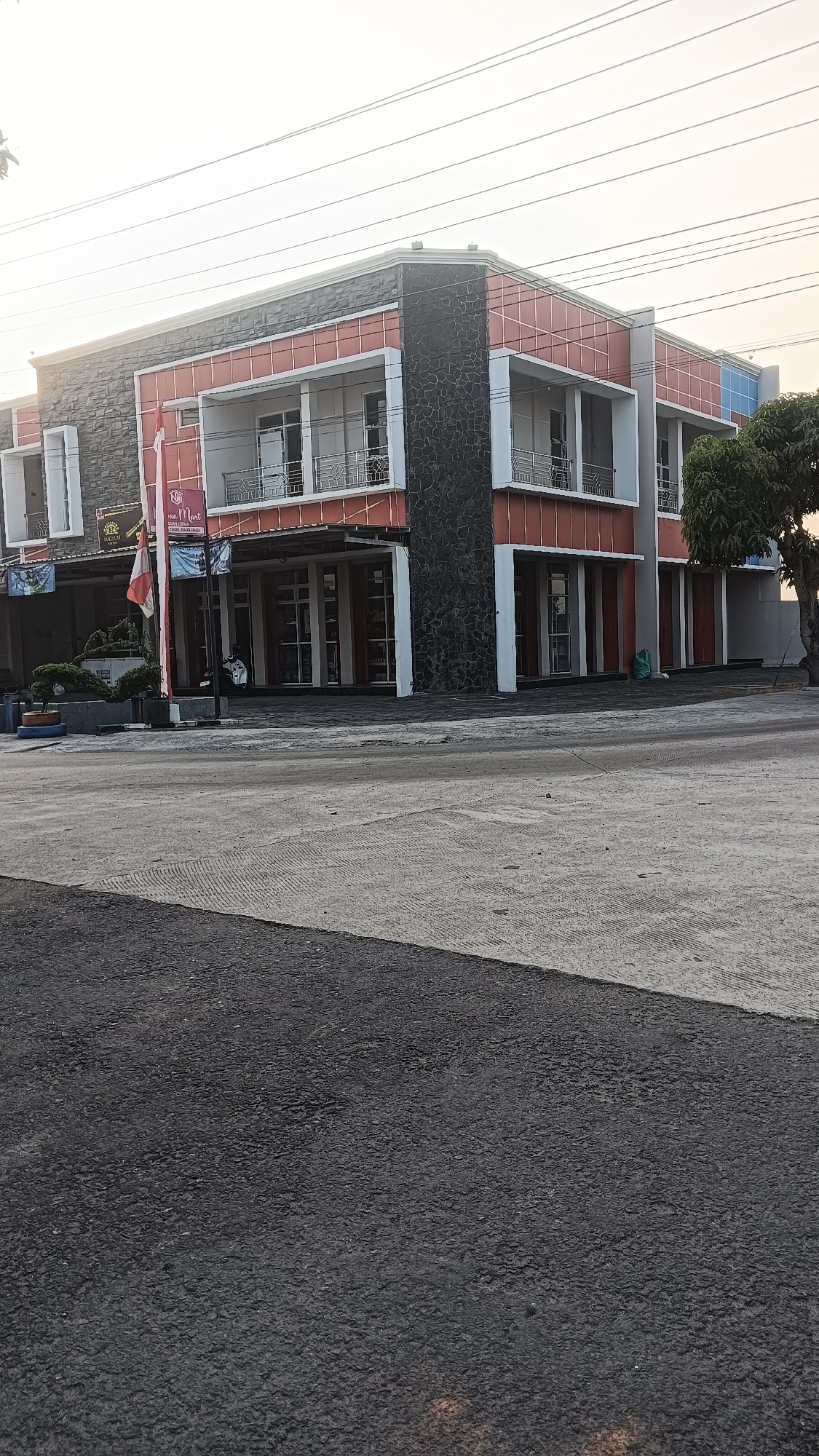 A two-story building with a modern design is situated in Tangkil, Indonesia. The building features a combination of orange, grey stone, and white facade elements. The ground floor has a row of storefronts with large glass windows and wooden doors. Above the storefronts, the second story has balconies with metal railings. A small tree with dark green leaves is visible on the right side of the building. In front of the building, there is a paved area with a street in the foreground. A red and white flag flies on a pole near the entrance of the building. Electrical wires are strung across the sky above the building. The sky is a light, overcast white.
