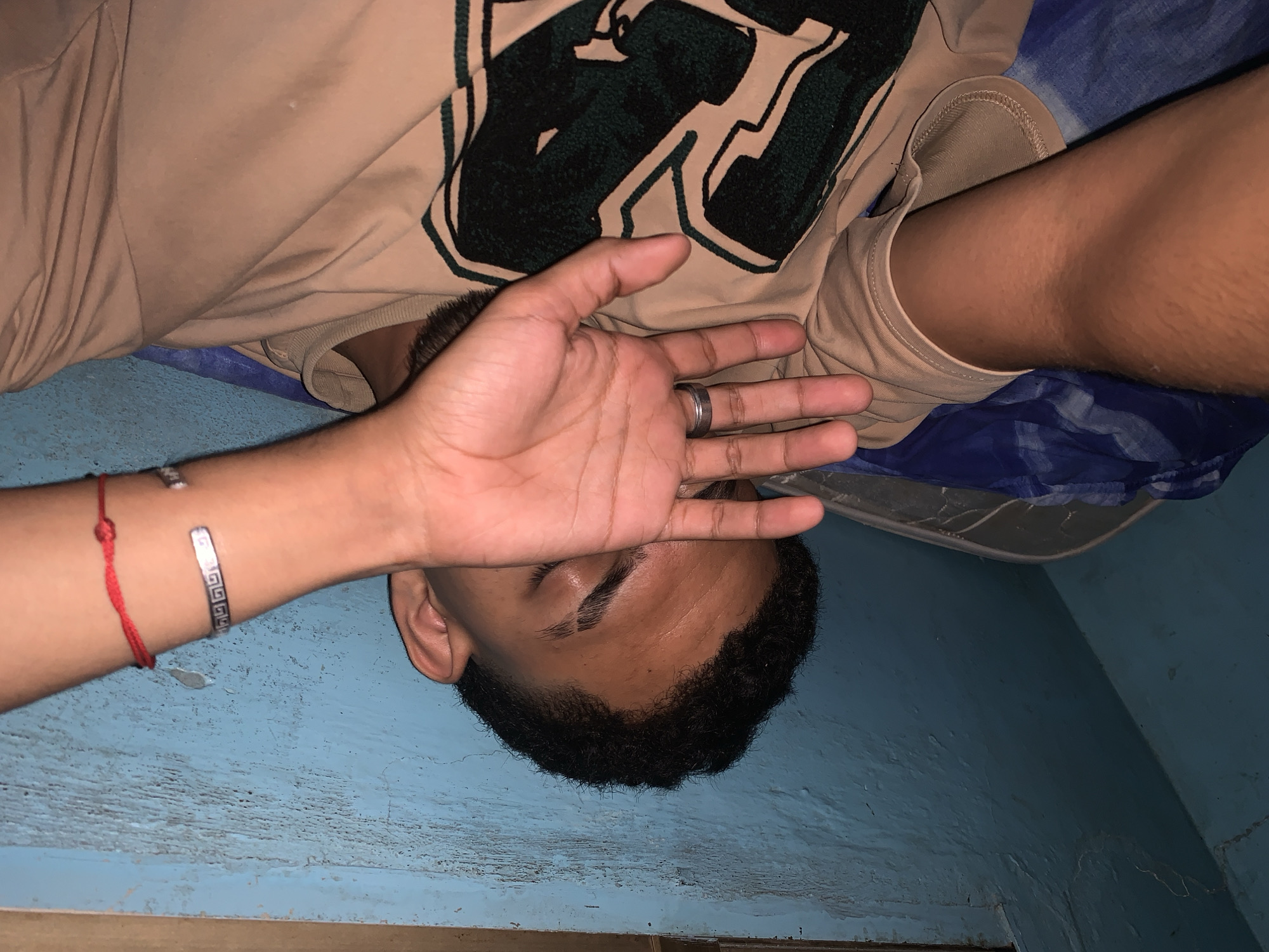 An overhead shot captures a person, likely male, lying down with their head oriented towards the bottom of the frame, in what appears to be an indoor setting in Santa Ana, Venezuela. The person's face is partially obscured by their left hand, which covers their eyes and forehead, suggesting rest or contemplation. They have dark skin, short dark hair, and are wearing a light brown (tan) t-shirt adorned with a prominent dark green, stylized graphic design on the chest, possibly a large block letter. The collar of a lighter-colored undershirt is visible beneath.
The person's left hand sports a silver-toned ring on the ring finger. Their left wrist is accessorized with two bracelets: a simple red string bracelet and a silver-toned cuff bracelet featuring a repeating Greek key pattern. The background consists primarily of a textured, light blue-painted surface, possibly a wall or floor, and a blue and white patterned fabric, likely a blanket or sheet, in the upper right portion of the image. The lighting is dim, contributing to a subdued atmosphere. No explicit text is legible beyond the stylized graphic and the bracelet pattern.