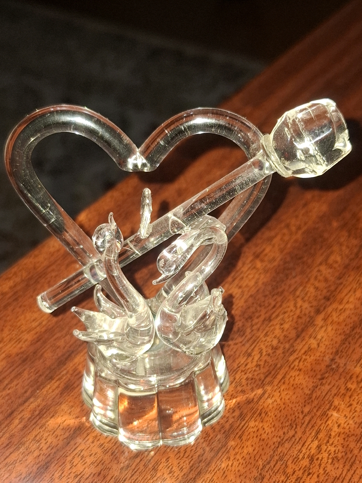 The image shows a glass figurine of two swans forming a heart shape with a rose. The figurine is placed on a polished wooden surface, likely a table. The background is dark and out of focus. The lighting suggests an indoor environment, possibly a home or a shop. There are no people or visible actions in the image. The scene depicts a decorative object. No information is provided to determine the location in Almaty, Kazakhstan.