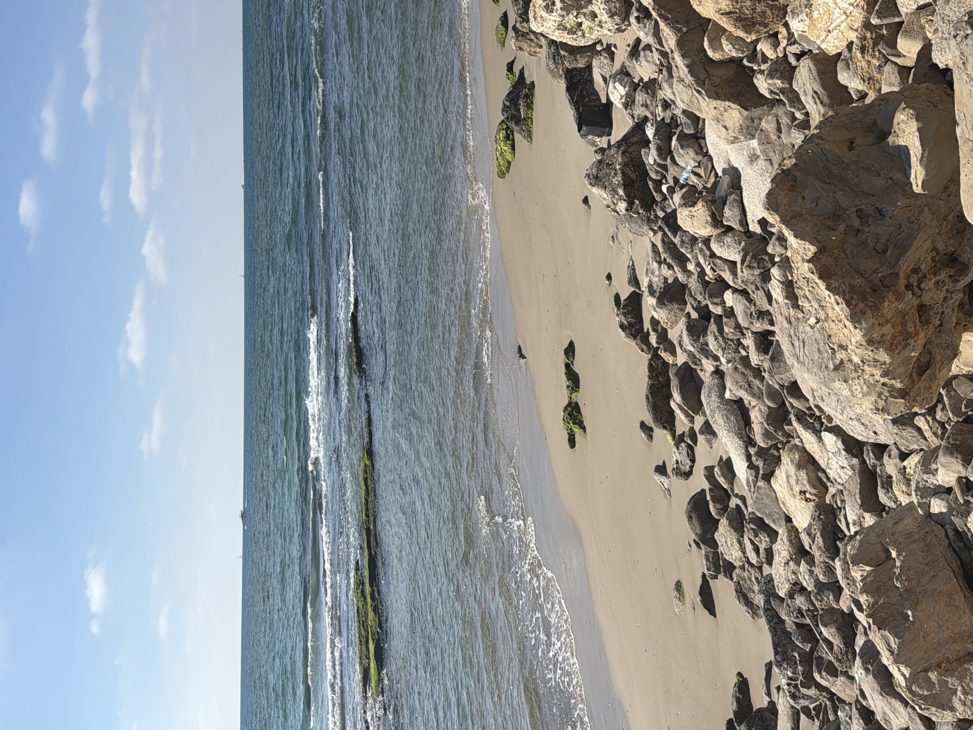 The image depicts a scenic view of the Tel Aviv coastline in Israel. The foreground is dominated by a rocky shore with large, weathered boulders of various sizes and textures. Some of these rocks are covered with green algae, suggesting their proximity to the water. A sandy beach stretches out behind the rocks, meeting the gentle waves of the Mediterranean Sea. The water is a clear, deep blue, with white foam cresting as waves approach the shore. In the middle ground, several submerged rocks or breakwaters, also covered in green algae, are visible just beneath the water's surface. The horizon is marked by the vast expanse of the sea meeting a pale blue sky dotted with scattered white clouds. Two small boats are visible in the distance, one appearing to be a sailboat and the other a motorboat. The overall impression is one of a serene and natural coastal landscape.