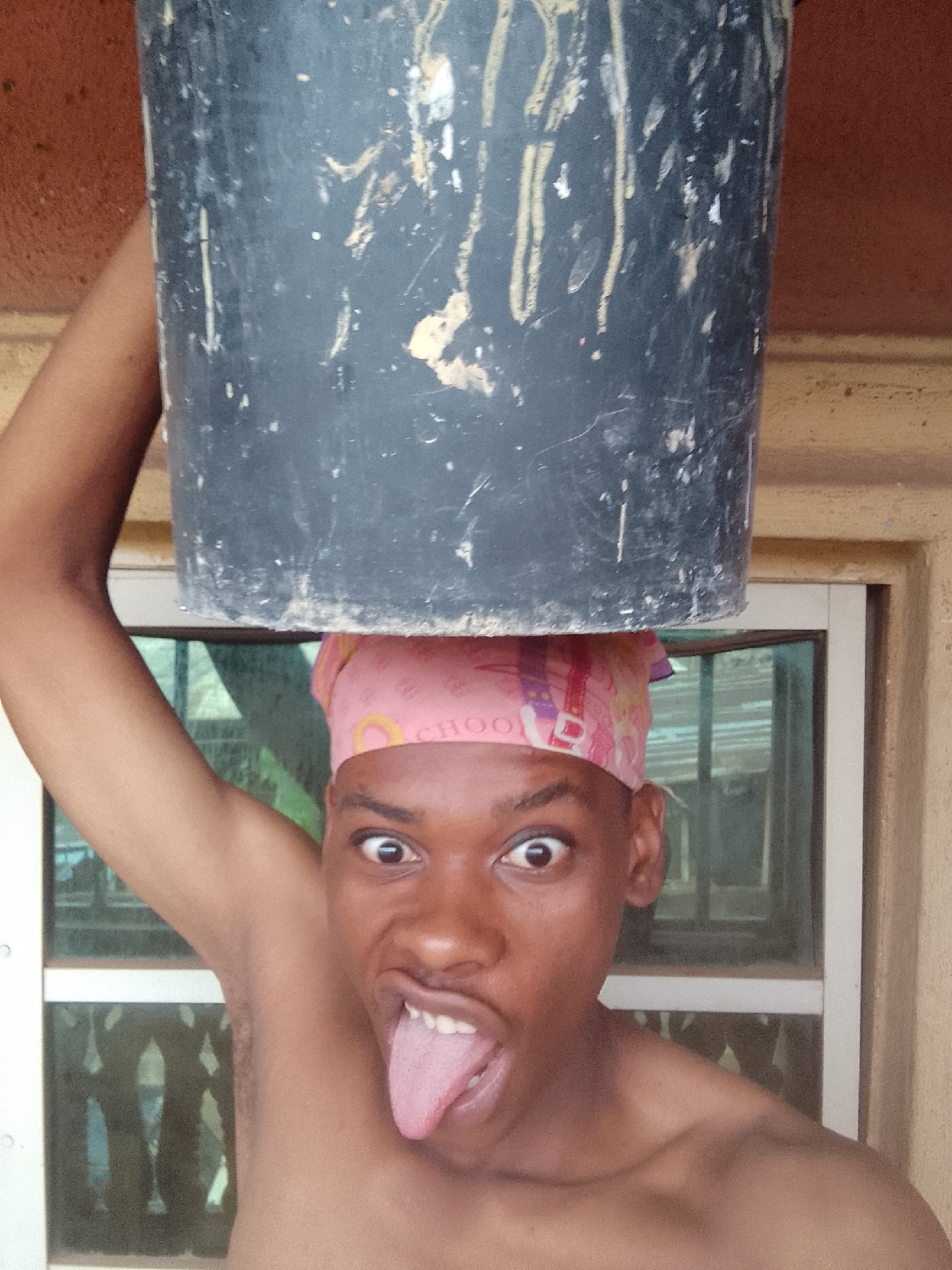A shirtless individual with a pink head covering is holding a black bucket on their head and sticking out their tongue. They are standing indoors in front of a window. The location is unknown.