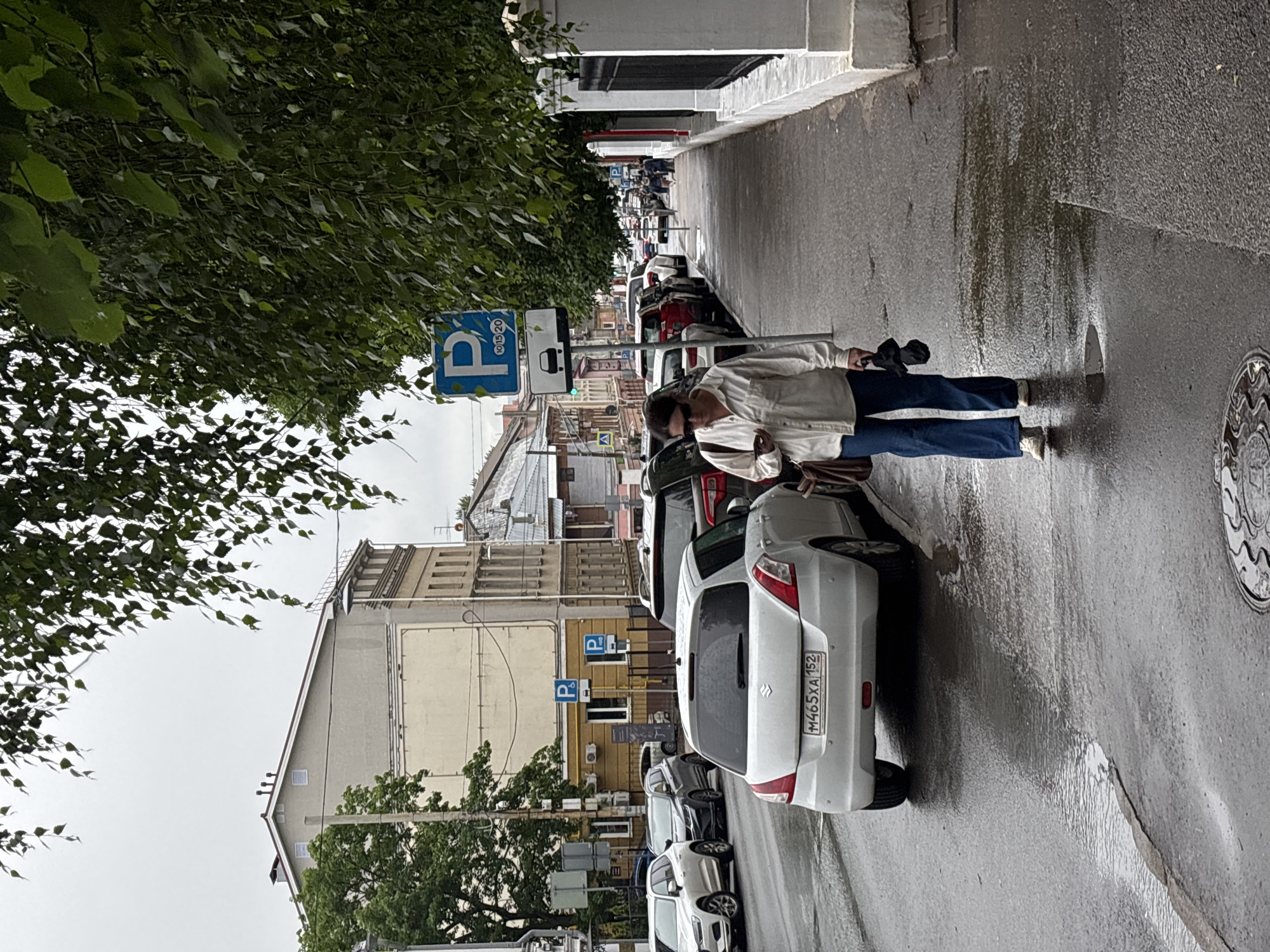 A person is walking down a wet city street in Nizhny Novgorod, Russia. The person, dressed in a light-colored jacket and dark pants, holds a black umbrella. They are walking on a sidewalk adjacent to a row of parked cars. The street is paved and appears to have recently rained, with visible puddles and wet patches. Buildings line the other side of the street, featuring architectural styles common to urban Russia, with windows and some signage. A prominent blue parking sign with a "P" is visible on a pole, along with other traffic signs. Lush green trees overhang the sidewalk from the left side of the frame, suggesting a temperate climate. The sky is overcast, indicating a cloudy or possibly rainy day. The overall impression is one of a typical urban scene on a damp day. Visible text includes a license plate on one of the cars, "M465XA 152".