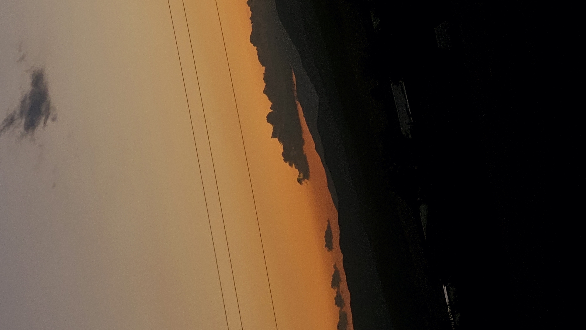 A vertical, eye-level shot captures a dark silhouette of rolling hills against a gradient orange sky, indicative of a sunset or sunrise. Several thin, horizontal power lines stretch across the upper portion of the frame, partially obscuring the sky. Small, dark clouds are scattered across the horizon, with a more prominent cluster of clouds casting a shadow over the upper slopes of the hills. The lower portion of the image is almost entirely black, making it difficult to discern any details of the landscape or any structures present. The image appears to be taken outdoors in a rural or semi-rural setting.