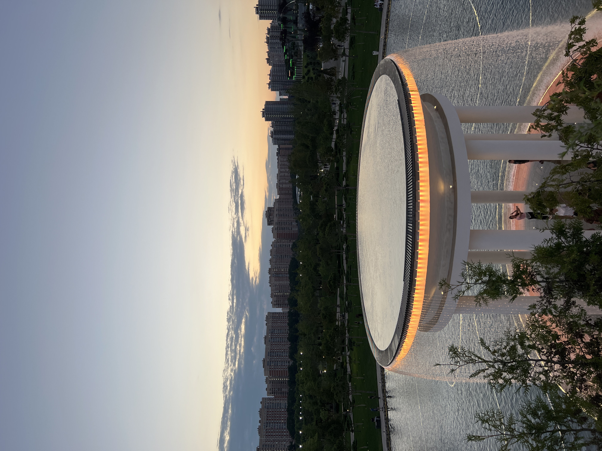 This image depicts a serene urban park scene at dusk, likely in Krasnodar, Russia. The focal point is a large, circular fountain with a prominent rim illuminated by warm, orange lights. Water cascades from the fountain, creating a shimmering effect on the surface.
In the background, a row of modern apartment buildings stretches across the landscape, interspersed with lush green trees and well-maintained lawns. The sky transitions from a clear, pale blue on the left to a soft, golden hue on the right, indicative of sunset. Wisps of clouds add texture to the twilight sky.
Several people are visible in the foreground and midground, some casually strolling and others observing the fountain. Their presence suggests a public space that is frequented and enjoyed by residents. The overall mood is peaceful and relaxing, enhanced by the soft lighting and the gentle sounds of the water feature. The time of day is clearly evening, with the natural light fading and artificial lights beginning to take prominence. The setting appears to be a well-designed urban park or public square, designed for recreation and aesthetic appeal. No visible text is present in the image.