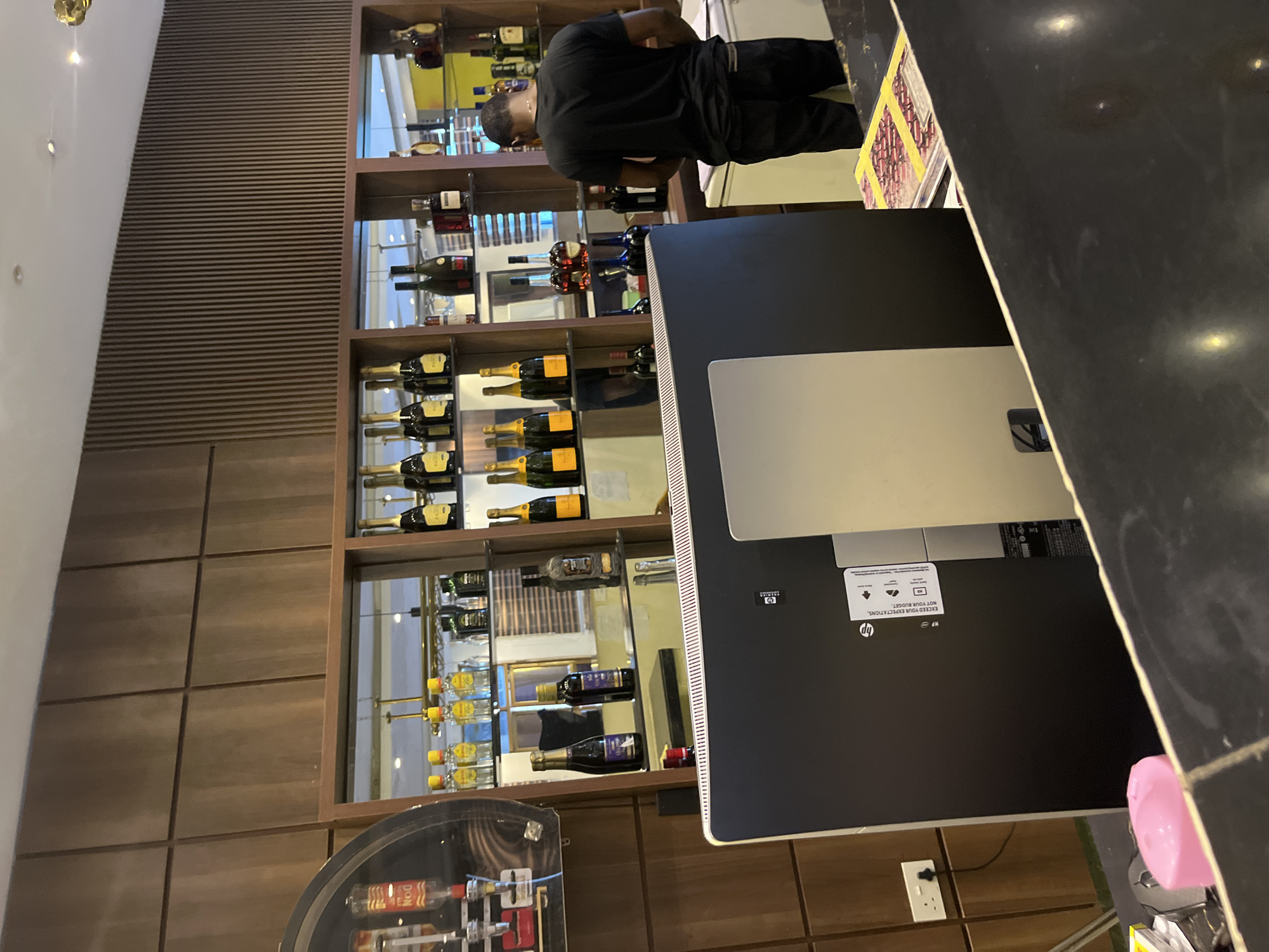 The image shows the interior of a bar or lounge, likely in Abuja, Nigeria. A man in a black t-shirt and black pants stands behind a counter, looking down at a piece of paper. In front of him is a large, black computer monitor. The background features shelves stocked with various bottles of alcohol, including champagne and spirits. The bar area is well-lit and appears to be modern in design, with wooden paneling on the walls. The overall atmosphere suggests a commercial establishment.