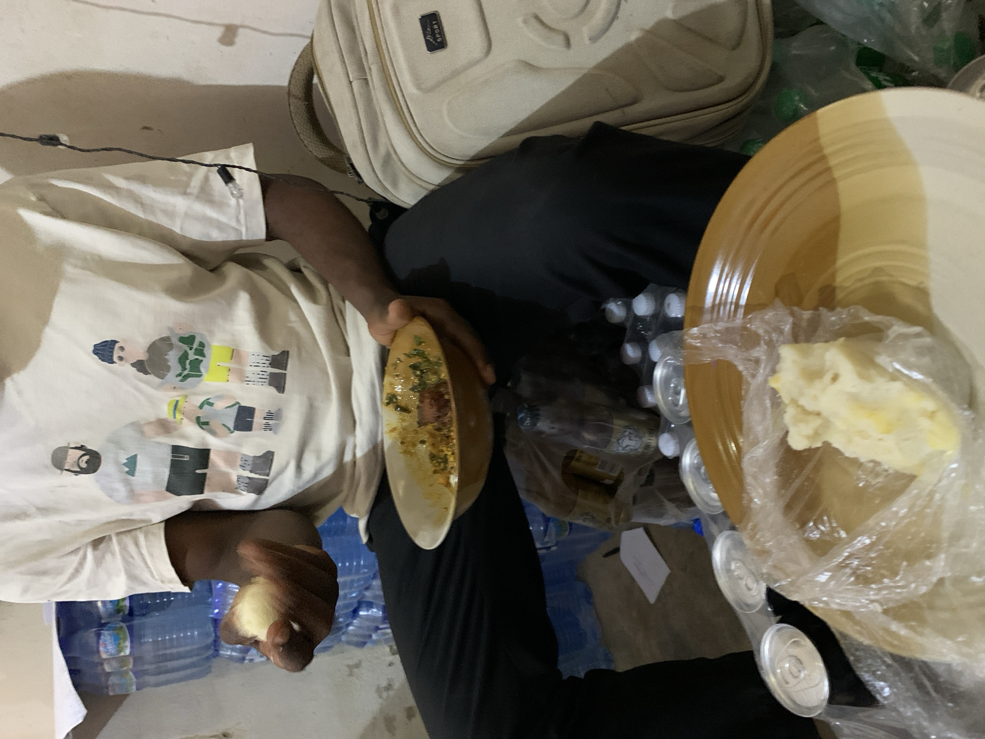 A person with dark skin is holding a plate with food and a wrapped portion of food, in what seems to be an indoor setting. The individual is wearing a white t-shirt with a cartoon-like design and black pants. Beside them is a light-colored backpack with a logo. Several bottles of water and cans are scattered around, suggesting a possible gathering or event. The location is referenced to be Kabong, Nigeria.