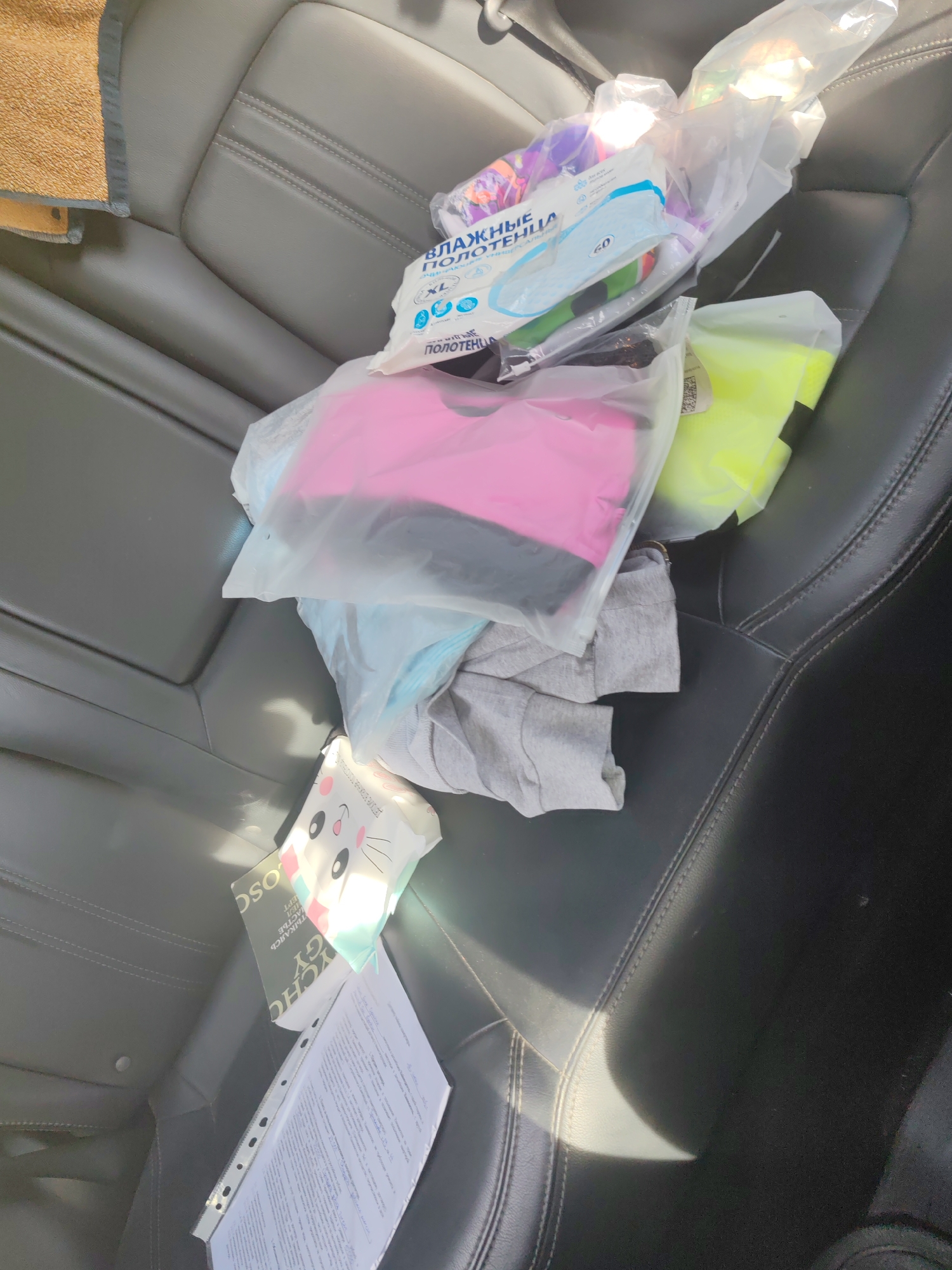 The image shows the interior of a car, likely a backseat, with various items scattered on the seats. A brown towel is visible on the left side. Several plastic-wrapped items are present, including a package of wet wipes with Russian text that reads "ВЛАЖНЫЕ ПОЛОТЕНЦА." There are also brightly colored items, one pink and one neon yellow, inside plastic bags. A grey t-shirt is partially visible. Additionally, a book with Russian text and a binder containing papers are on the seat. The car's interior appears to be made of black leather. The lighting suggests it is daytime, with sunlight casting shadows. The specific location in Paratunka, Russia is not discernible from the image itself; this context would have been provided externally.
