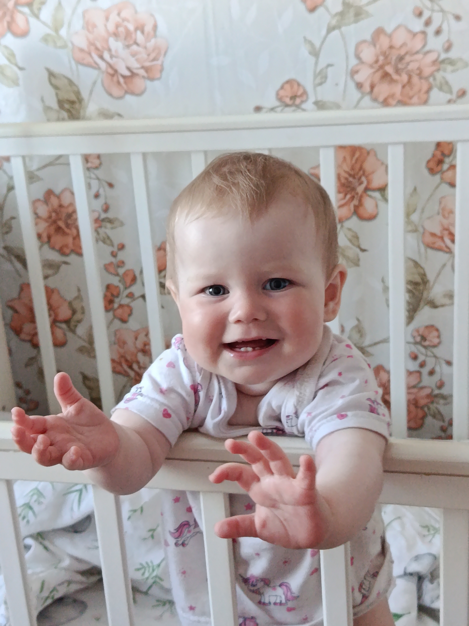 A baby is standing in a white crib with floral wallpaper in the background. The baby has fair skin, blonde hair, and blue eyes. The baby is wearing a white outfit with small pink hearts and unicorn prints. The baby is smiling with two visible teeth and holding their hands up. A patterned sheet is visible inside the crib. The location is Фоки, Russia.