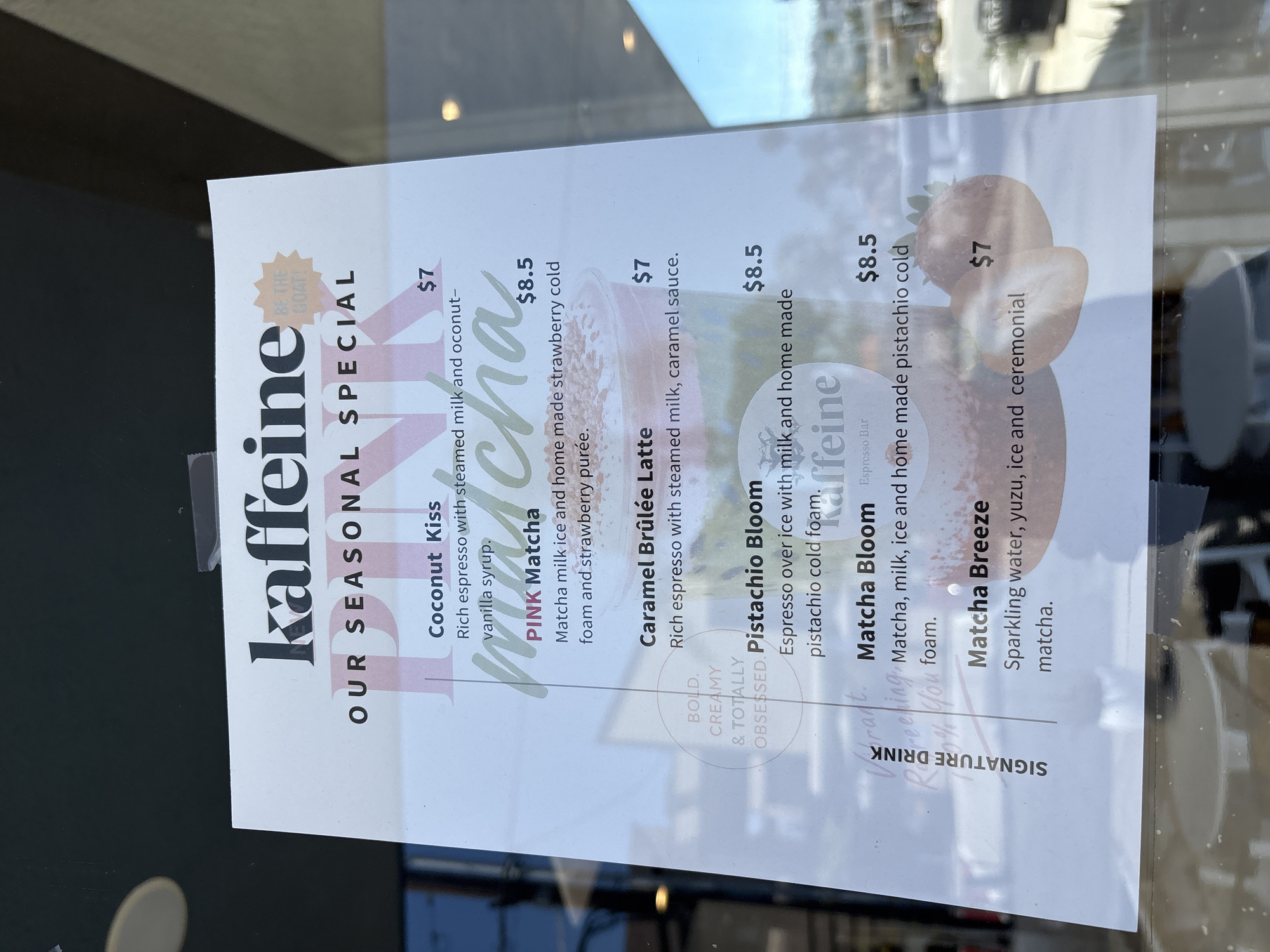 Here is a forensic description of the image.
The image shows a sign advertising seasonal specials at a café named "Kaffeine." The sign is taped to a window, likely of the café itself. The sign is white with black and pink text. The specials listed are:
- Coconut Kiss ($7)
- PINK Matcha ($8.5)
- Caramel Brûlée Latte ($7)
- Pistachio Bloom ($8.5)
- Matcha Bloom ($8.5)
- Matcha Breeze ($7)
The lower part of the sign reads "SIGNATURE DRINK." The text "BOLD, CREAMY & TOTALLY OBSESSED" is written in a semi-circular fashion on the left side of the sign. A graphic of a strawberry is visible at the bottom right of the sign.
The sign includes an endorsement indicating something or someone is 'the Goat!'
Based on publicly available information, there is a cafe named Kaffeine located in Redondo Beach, California, United States. The sign likely advertises specials at the Kaffeine located in Redondo Beach, California.