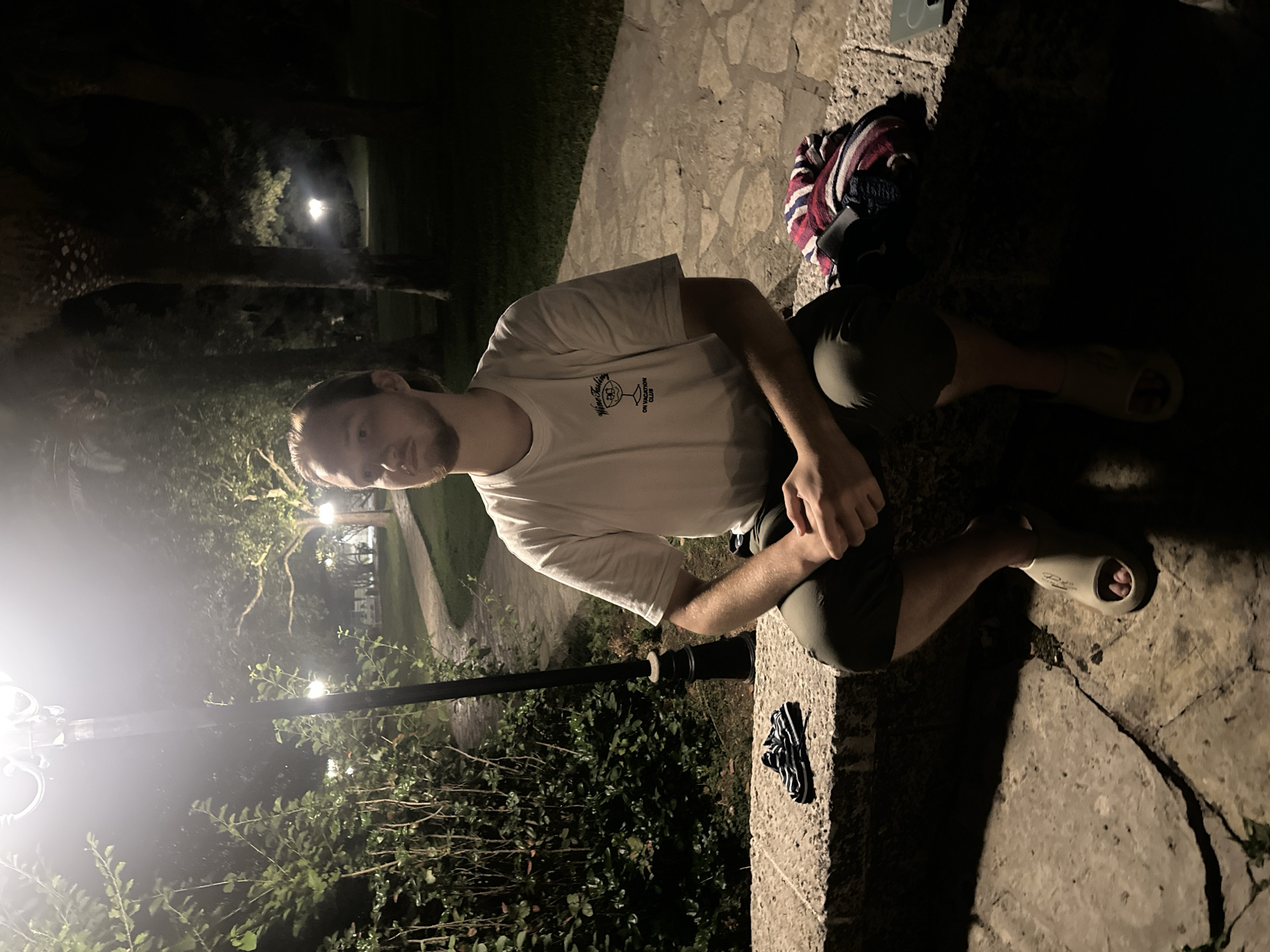 A young man with light brown hair and a beard sits on a stone ledge at night. He is wearing a white t-shirt with a graphic print and olive green shorts, along with beige slides. His hands are clasped in his lap as he looks to his left. Beside him on the ledge are a pair of black and white striped flip-flops and a folded blanket with red, white, and blue stripes. The background shows a pathway winding through a garden with trees and illuminated by streetlights. The scene appears to be in the vicinity of Budva, Montenegro, as indicated by the architecture and vegetation characteristic of the region.