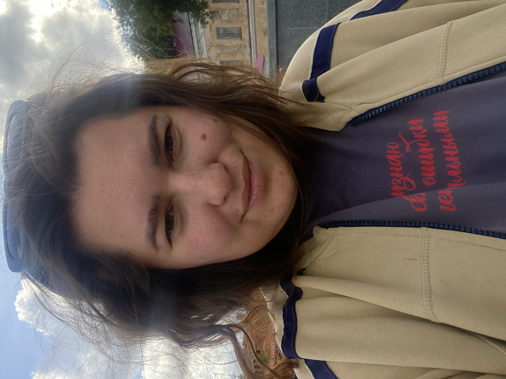 A young woman with medium-length brown hair is taking a selfie outdoors on a cloudy day. She is wearing a light beige zip-up jacket with blue stripes and a dark purple t-shirt with red text. The text on her shirt reads, "Я знаю ошибки плывущими" which translates to "I know mistakes are floating." She has freckles on her cheeks and a mole on her right cheek. Behind her, a yellow building with windows and a red roof is visible, along with some trees and a cloudy sky. The location is the Perm State Art Gallery in Perm, Russia, recognizable by the building's architecture.