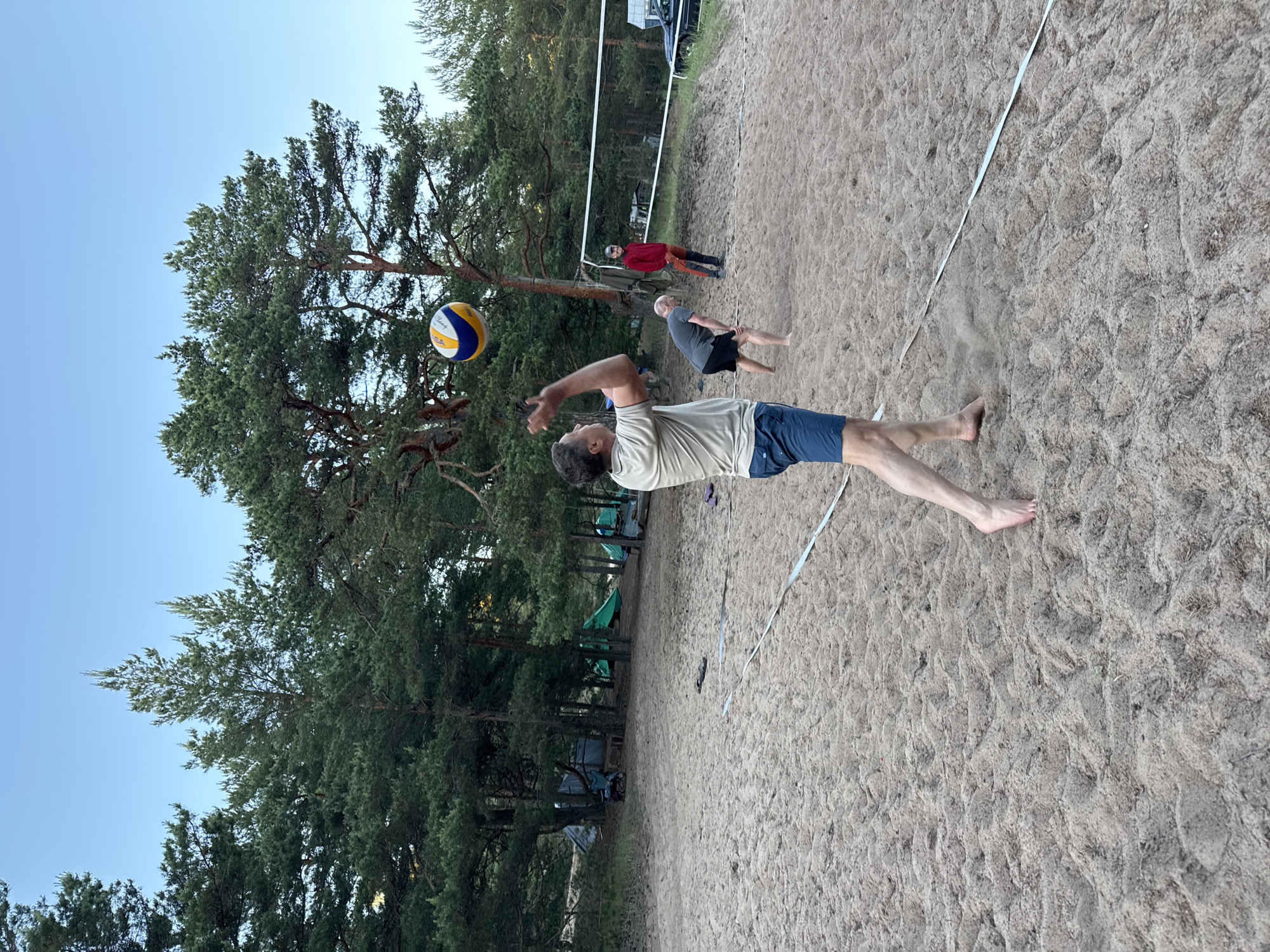 In Zelenaya Roshcha, Russia, on a sandy beach volleyball court, a man in a light brown t-shirt and blue shorts is captured mid-action, preparing to serve a volleyball. His body is angled, and his arms are raised, with the ball in the air above his head. Another man in a grey t-shirt and black shorts crouches on the court, appearing to be in a defensive stance. A third man, wearing a red hooded jacket and orange pants, stands further back on the court. In the background, a volleyball net stretches across the sand, with pine trees and several tents set up amongst them. The sky above is a clear, pale blue, suggesting it is either early morning or late evening. The sandy ground is imprinted with footprints and the texture of the sand is clearly visible.