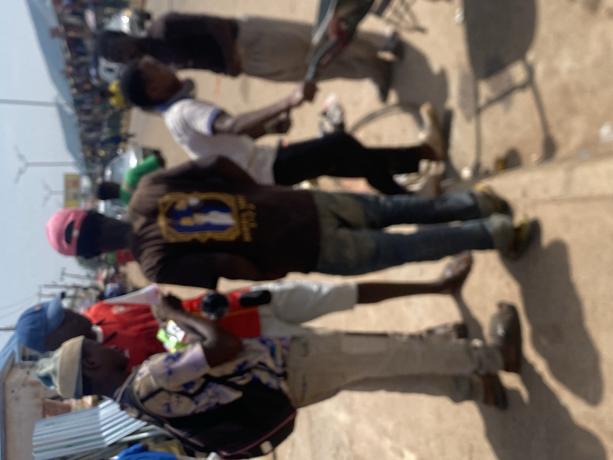 In Bukuru, Nigeria, a group of individuals is gathered on a dirt road, appearing to be a market or busy street. The scene is slightly blurred due to motion or focus issues. In the foreground, a man wearing a patterned shirt and light-colored pants is looking down at an object in his hands, possibly a phone or small item. Beside him, another person in a red shirt and white shorts stands with a blue baseball cap. Further back, a person in a dark t-shirt with a graphic on it, and another in a white t-shirt with black pants, are visible. The background shows a street with several cars, some buildings, and many people browsing stalls, suggesting a bustling commercial area. A wheelbarrow is also present, indicating potential trade or transport activities. The overall impression is a snapshot of daily life in a Nigerian urban setting.