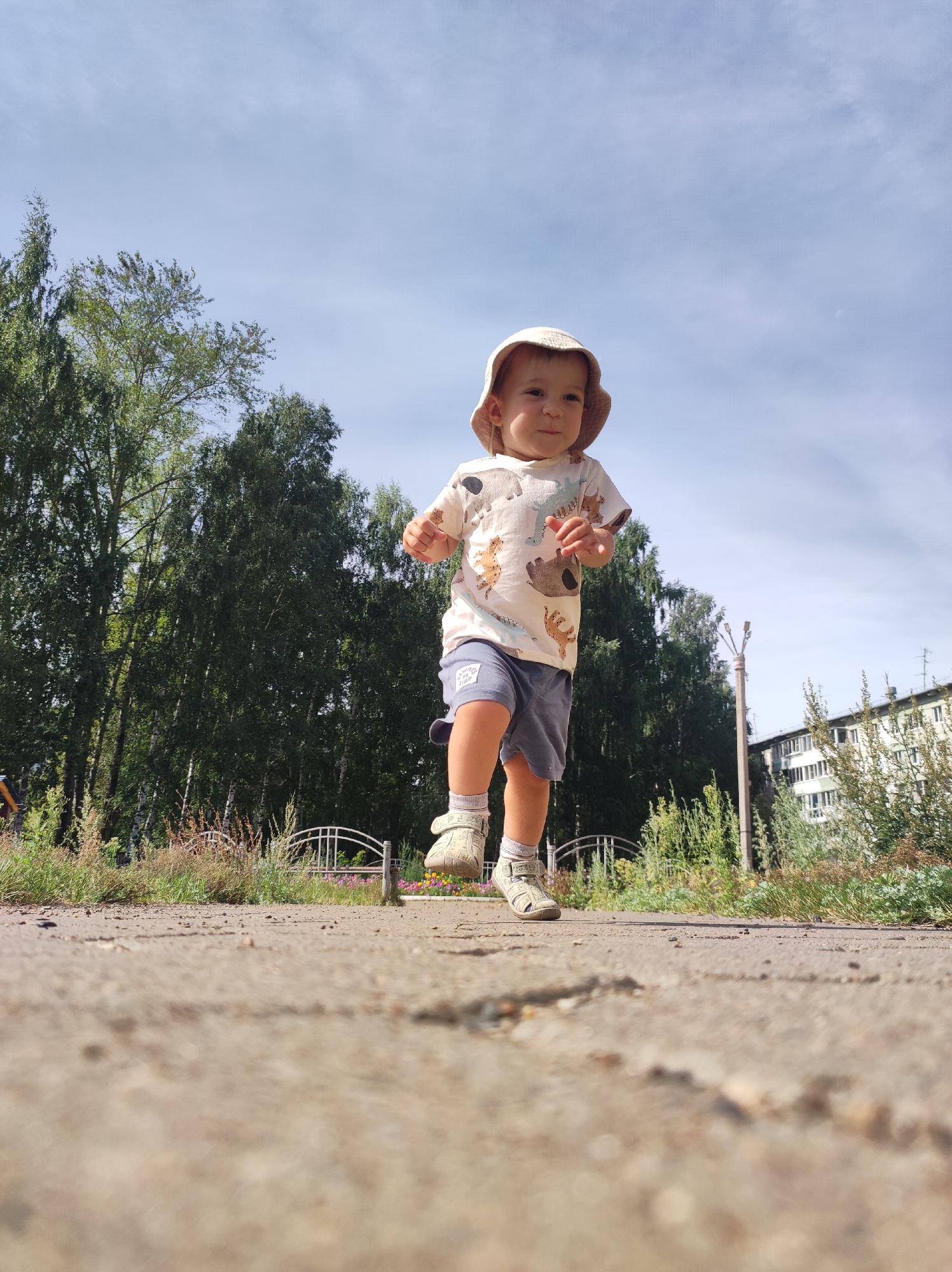 A low-angle, full-shot photograph captures a toddler running towards the camera on a concrete path. The child, wearing a light-colored bucket hat, a t-shirt with animal prints, and gray shorts, is positioned in the center of the frame, mid-stride. The background features a line of tall, green trees, a white metal fence, and a multi-story apartment building in the distance, suggesting an urban or suburban park setting. The sky is partly cloudy and a bright blue. The image appears to have been taken in Chaikovsky, Russia, given the presence of birch trees common to the region and the architectural style of the building. The foreground is dominated by the textured concrete path, with some scattered pebbles.