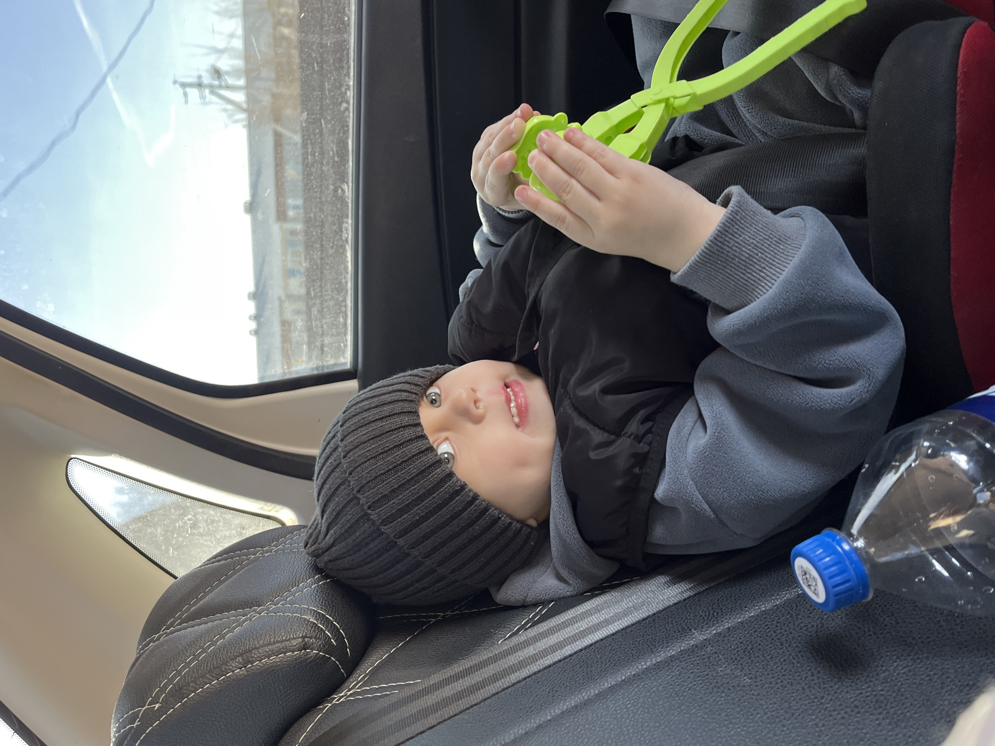 A young child wearing a gray knit hat, a gray hooded sweatshirt, and a black vest is sitting in a black car seat, holding a green plastic object. The car seat is positioned in the back of a vehicle, and a partially empty plastic water bottle with a blue cap is visible. Outside the car window, a building and a utility pole are partially visible, suggesting an urban or suburban setting. The child is looking upward with a slight smile. This is likely not in Фоки, Russia since the language on the bottle cap is English.
