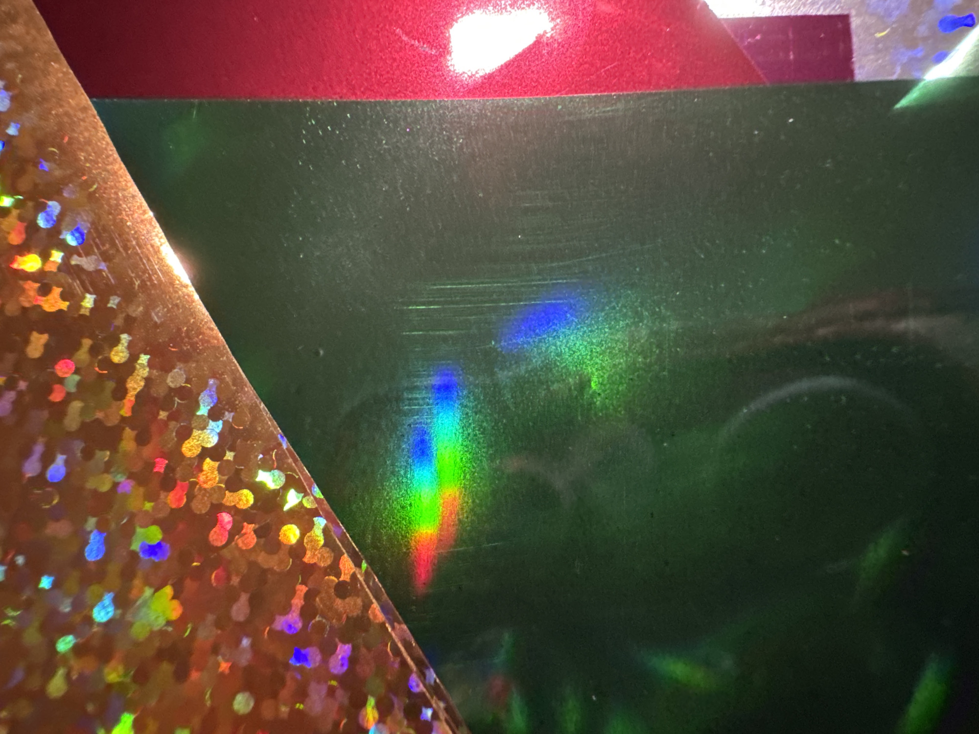 The image shows a close-up, abstract view of various textured surfaces with holographic and metallic sheens. In the foreground, a dark green, metallic surface is visible, displaying faint scratches and reflections. A vibrant band of rainbow light, likely a reflection from a holographic source, is spread across this green surface. To the right of the green surface, a bright red, metallic surface is seen, with a strong white reflection on its surface. At the top of the image, a rose-gold colored surface with a distinct holographic pattern of small, irregular shapes is present, also exhibiting a variety of iridescent colors. The composition is characterized by sharp angles where the surfaces meet, creating a layered effect. The image does not depict any recognizable landmarks or features that would indicate a specific location in Waterford, Ireland.