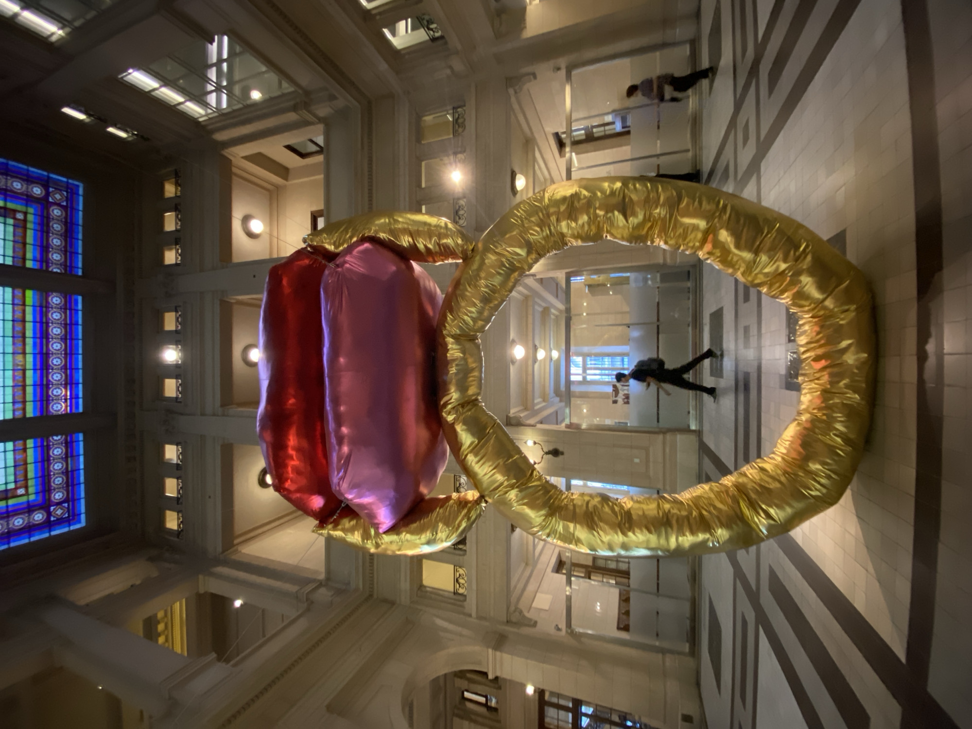 The image shows a large art installation resembling a ring with an oversized red and pink jewel in the center, suspended in the air. The ring itself is gold and appears to be made of an inflated material. The surrounding environment is a grand building interior, likely a public space, with multiple levels, arched windows, and ornate architectural details. There are people walking in the background on different floors. The perspective is tilted upwards, providing a unique view of the installation and the building's architecture. Given the presence of distinct architectural styles and decorative elements, it's speculated that the location is in the city of Buenos Aires, Argentina. The lighting is bright, illuminating the artwork and the interior of the building.