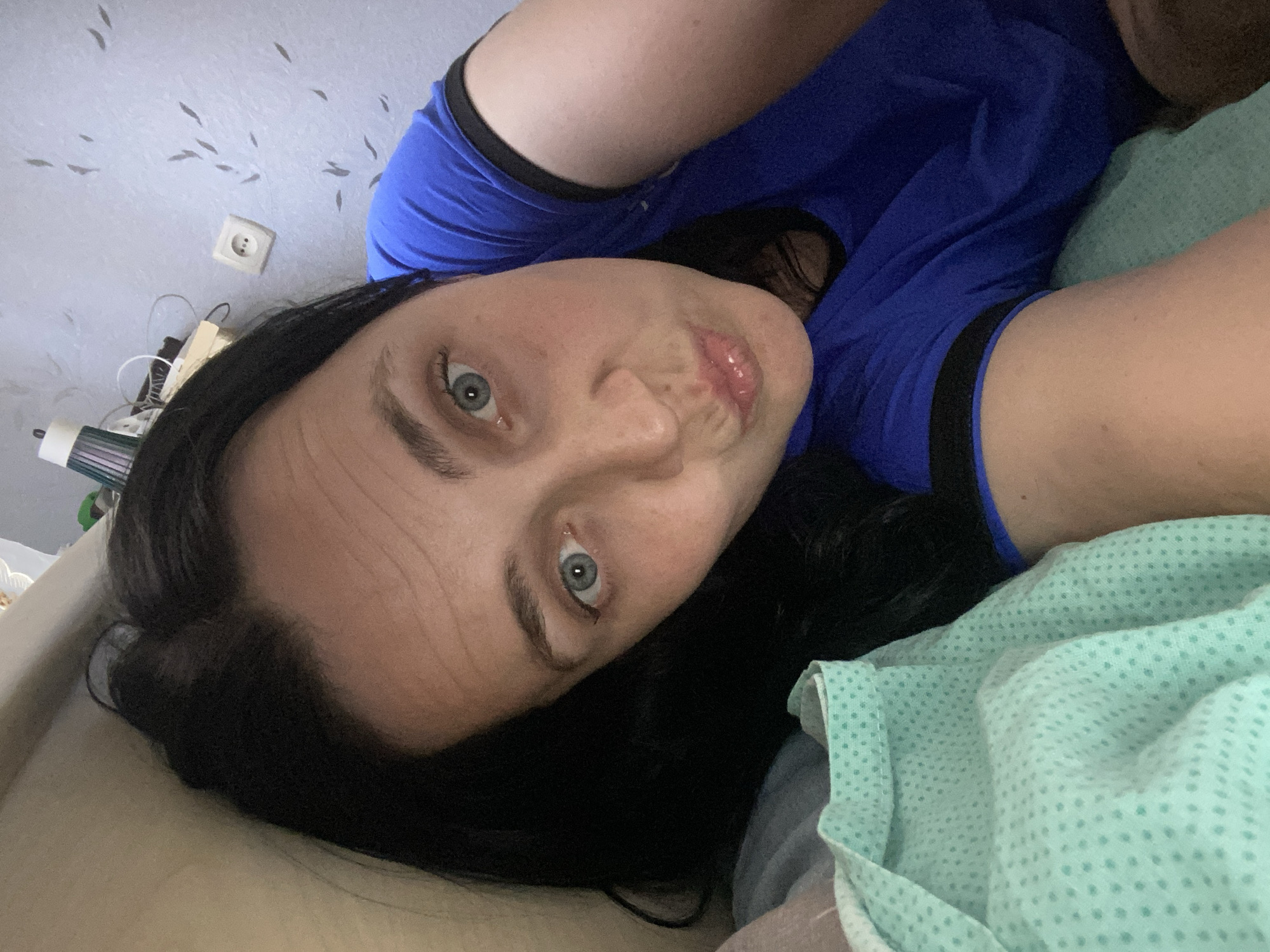 A woman with black hair and blue eyes is lying down, looking at the camera. She is wearing a blue t-shirt. She is in a bedroom with a light blue wallpaper with a pattern of small, dark gray shapes. There is a white electrical outlet on the wall. A light green patterned blanket is visible on the left side of the image. The overall lighting is soft and indoor.