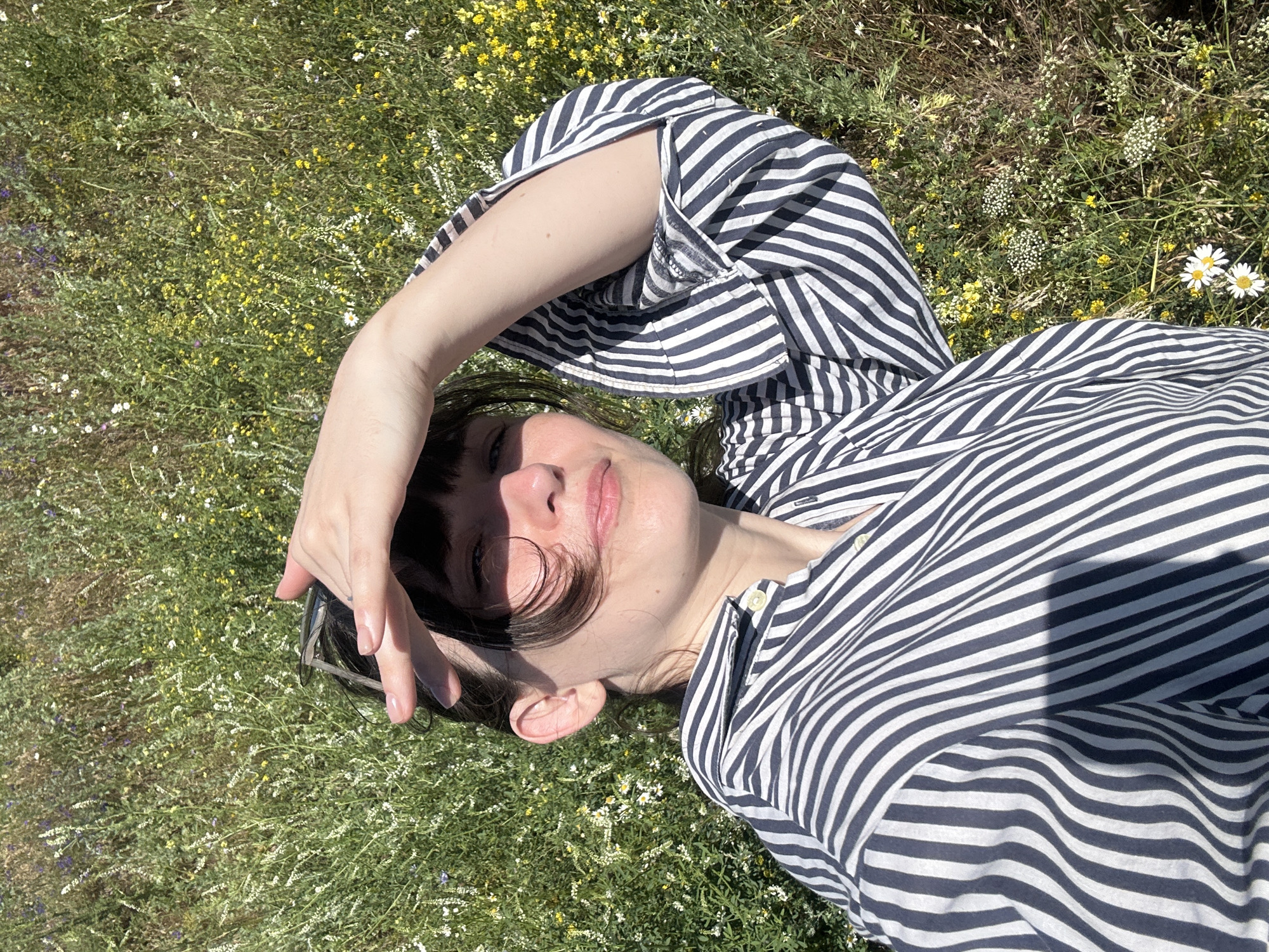 A woman is lying in a field of wild flowers, her head to the right of the frame. She is wearing a blue and white striped shirt and has her right hand shielding her eyes from the sun. Her left arm is bent and raised, with her hand resting on her forehead. The field is green and filled with small yellow and white flowers. The image was taken outdoors in Krasnyi Yar, Russia.