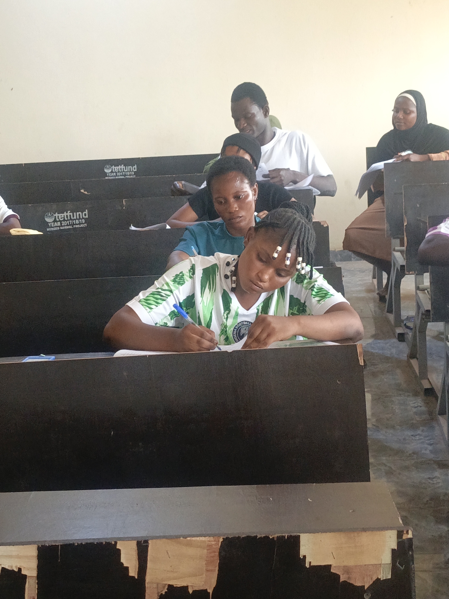 In an indoor setting, several individuals are seated at desks in what appears to be a lecture hall or classroom. The person in the foreground, wearing a white and green Nigeria Football Federation jersey, is focused on writing, with a blue pen in hand, on a notebook placed on the desk. Behind her, another woman with short black hair is also seated at a desk. Further back, a man in a white shirt is visible, along with a woman wearing a hijab on the right side of the frame. The desks are dark brown with signs indicating "tetfund YEAR 2017/18/19 MERGED NORMAL PROJECT" on them. The walls are a light beige color. The lower part of the desks is visibly damaged.