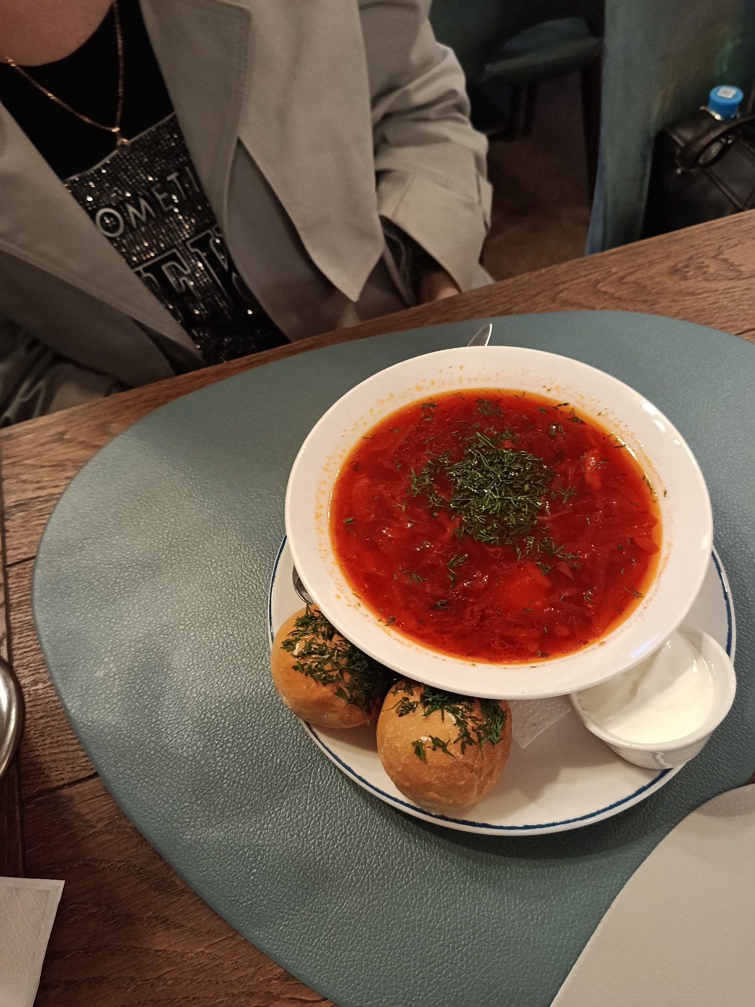 The image shows a person wearing a grey jacket and a black t-shirt with a silver sparkly design. On the wooden table in front of them is a blue-grey placemat. On the placemat, there's a white plate containing a bowl of red soup garnished with dill, two round bread rolls topped with herbs, and a small white bowl of sour cream. Part of another white plate is visible at the bottom right. There are hints of a green chair and a bag in the background. The location is Unknown, Unknown.