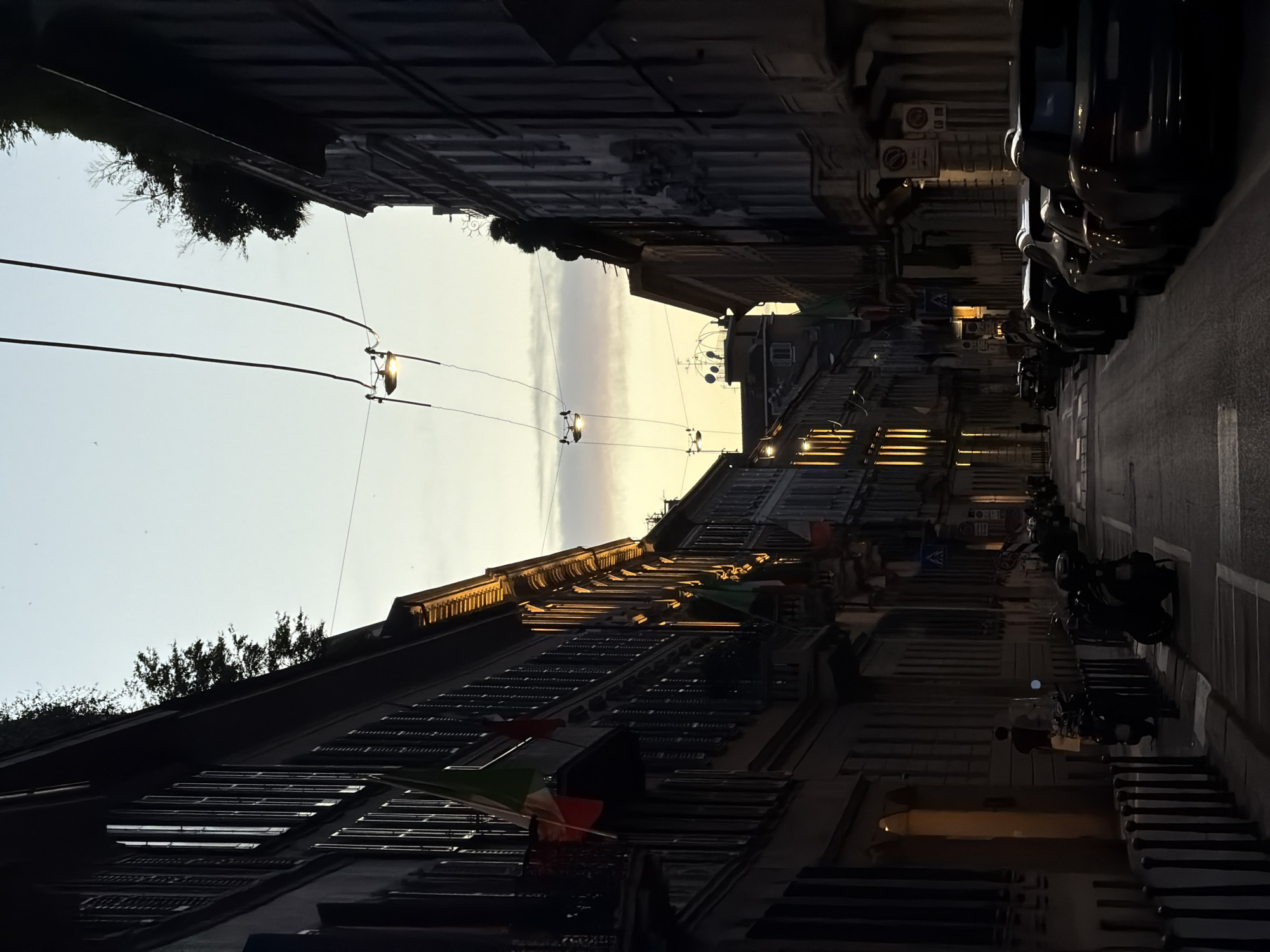 A street scene in Milan, Italy, captured during twilight. The image focuses on a narrow street lined with tall buildings on both sides. The buildings exhibit traditional architecture with shuttered windows and some ornate detailing. Italian flags are visible hanging from some of the balconies. Overhead, electrical wires crisscross the sky, supporting suspended streetlights that cast a warm glow on the street below. The road is paved and appears to be a one-way street, with a line of parked cars on the right side and a row of scooters and motorcycles parked on the left. In the distance, the street recedes, lined with more buildings. The sky above is a gradient of pale blue and soft orange, indicating the transition from day to night. A single person is visible on the sidewalk on the left side of the street, walking away from the camera. The overall atmosphere is serene and urban.