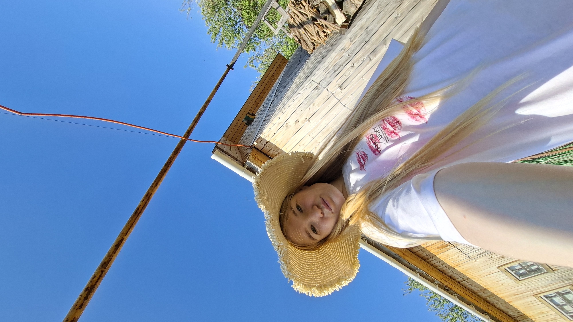 The image shows a low-angle view of a young woman in Большой Букор, Russia. She is wearing a straw hat and a white t-shirt with a design that includes the phrase "I am so lucky" and red lipstick kisses. She is lying on a wooden deck. Above her is a clear blue sky, and a rusty pole extends into the sky. The wooden building and pile of firewood are in the background.