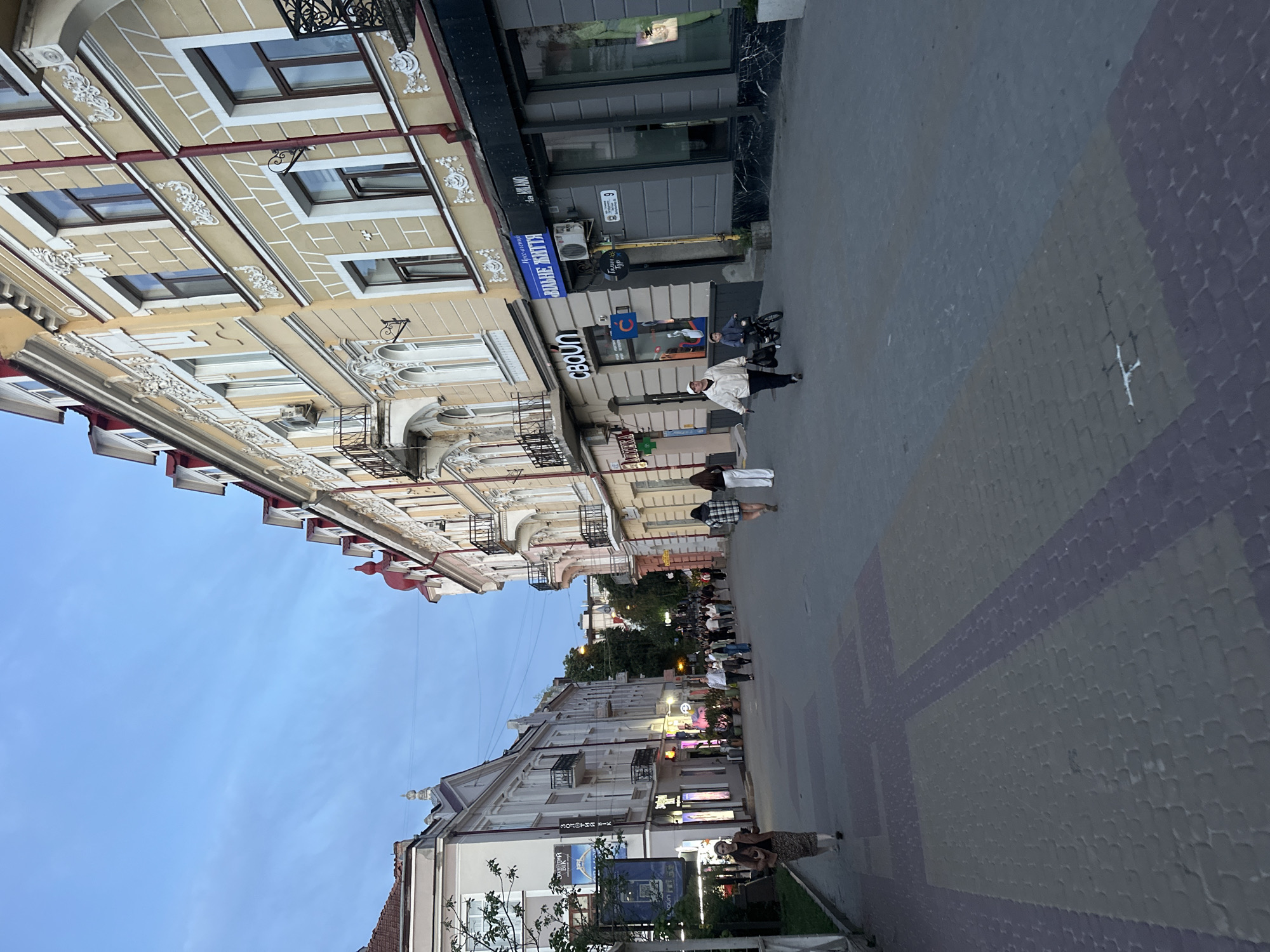 This image captures a view of Valova Street (Валова вулиця) in Ternopil, Ukraine, during what appears to be the early evening or late afternoon. The scene depicts a pedestrianized thoroughfare flanked by multi-story buildings with varied architectural styles.
On the right side of the street, a row of yellow-ochre colored buildings with white stucco decorative elements, including ornate window frames and cornices, extends into the distance. These buildings feature multiple dark-framed windows, some with attached black wrought-iron balconies. Red sloped roofs with dormer windows are visible at the top. Ground-level storefronts include businesses such as "СВОЇ" (SVOI), "ВІЛЬНЕ ЖИТТЯ" (Vilne Zhyttia - Free Life, a local newspaper), "Галицький Тур" (Halytskyj Tour - Galician Tour), and "Da MILANO." A street number "9" is prominently displayed on the building housing "ВІЛЬНЕ ЖИТТЯ." An outdoor air conditioning unit is visible on one building facade.
On the left side of the street, buildings are predominantly light grey or beige, also featuring decorative elements and balconies with black railings. Visible businesses on this side include "АТЕКА" (Apteka - Pharmacy), "ЗДОРОВИЙ ВІК" (Zdorovyi Vik - Healthy Age), and "ЗОЛОТИЙ ВІК" (Zolotyj Vik - Golden Age, a jewelry store). Green foliage from trees or shrubs is present along the left side of the street and in the distance.
The street itself is paved with interlocking pavers arranged in geometric patterns, primarily consisting of grey, lighter grey, and reddish-brown blocks. Numerous individuals are present on the street. In the midground on the right, a woman in a white jacket and dark pants walks alongside a young child riding a small blue bicycle, both moving towards the left. Further ahead on the right, a group of four women walks away from the camera. On the left, a woman in a patterned skirt and brown jacket walks away from the camera. A larger congregation of people is visible in the distance, some appearing to be seated at outdoor establishments. The sky above is a clear, light blue with subtle wisps of clouds, indicating ambient natural light, while some artificial lights from shops are beginning to illuminate. Overhead power lines are visible against the sky.