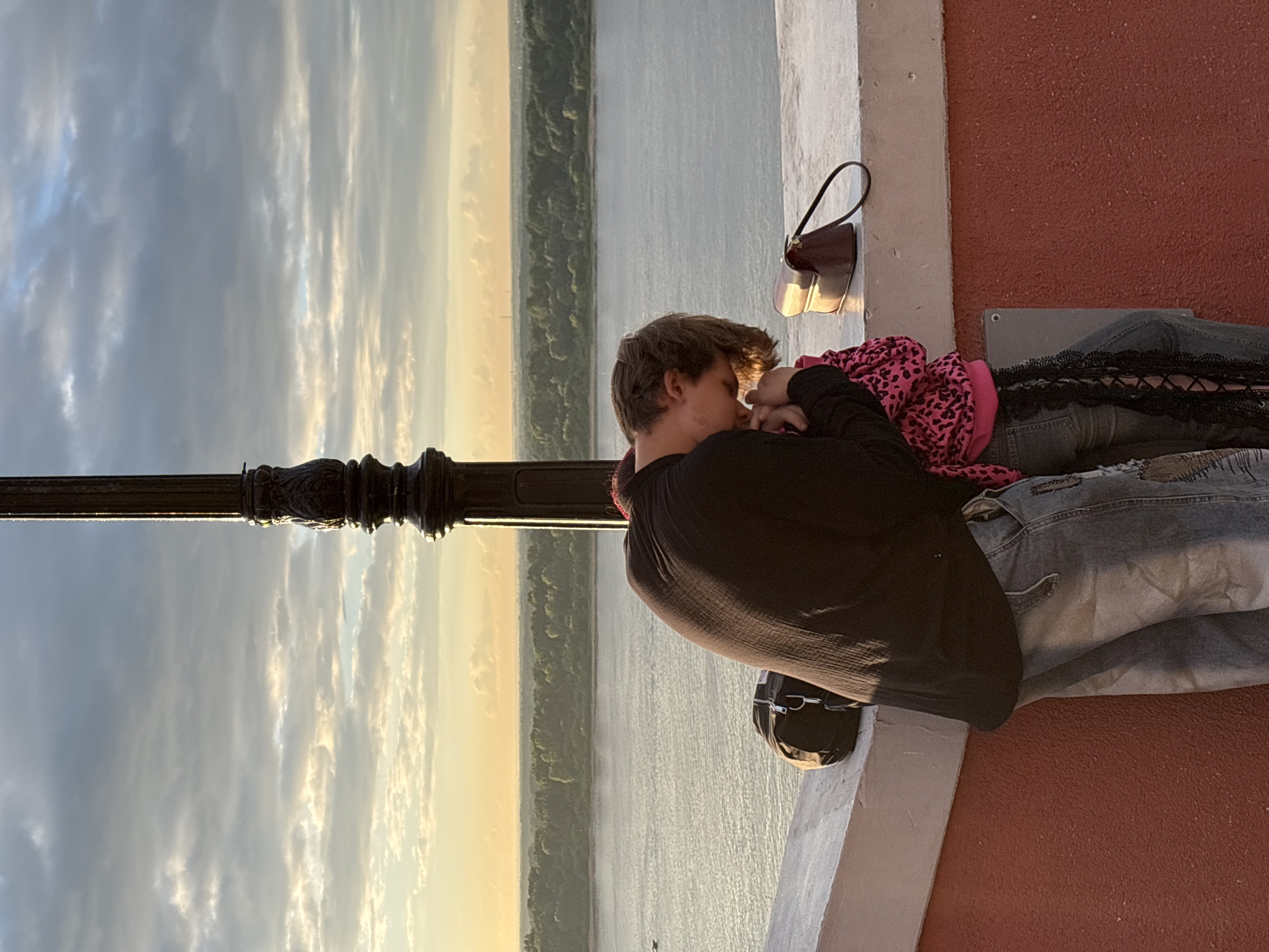 Two individuals, appearing to be a young man and a younger person, are embracing and kissing on a railing overlooking a wide river, likely the Volga River, in Nizhny Novgorod, Russia. The man, wearing a dark long-sleeved shirt and distressed denim jeans, is leaning down towards the other person. The other individual is wearing a pink leopard-print hooded sweatshirt over jeans with black lace detailing. A black duffel bag rests on the railing to the left of the couple, and a brown handbag is on the railing to the right. A decorative lamppost is positioned behind them, and the sky is filled with dramatic clouds at sunset. The background shows a tree-lined shore across the river. The ground beneath the railing is a textured red wall.