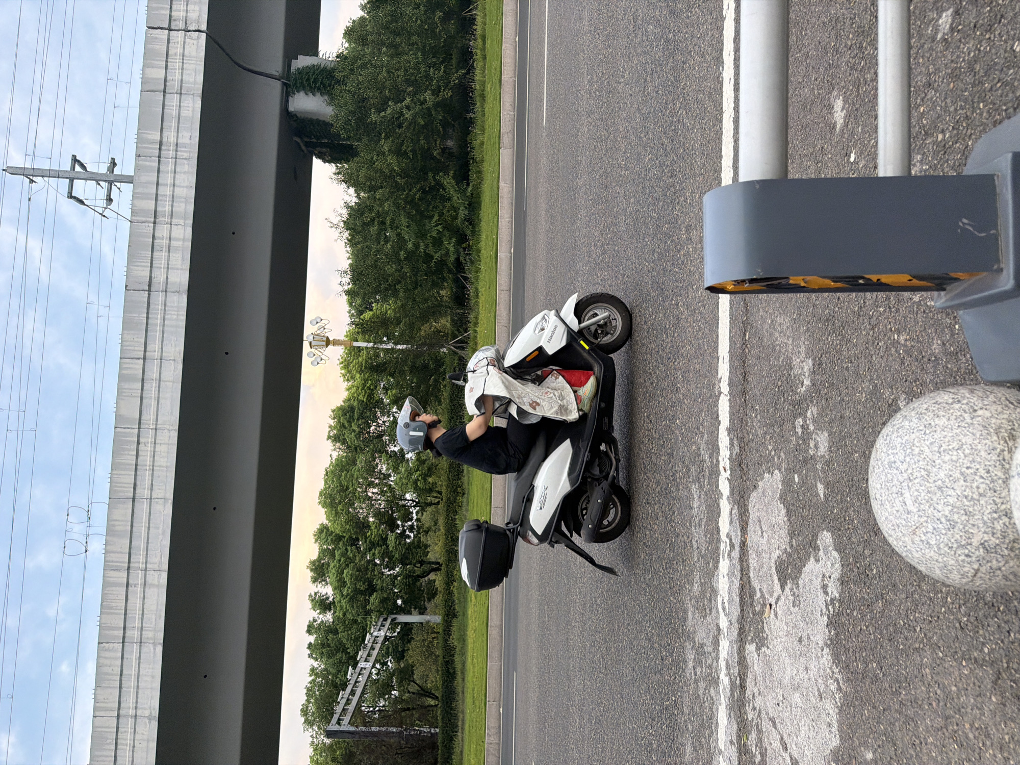 In Jiangjin, China, a person wearing a black shirt and a grey helmet rides a white scooter on a paved road. The scooter has a black storage box on the back. A white cloth with a floral pattern is draped over the front of the scooter. In the background, there is a large concrete overpass with power lines above it. Trees and greenery are visible on the side of the road, and a lamppost with multiple lights stands among them. The sky is cloudy. The foreground shows a gray bollard and part of a metal railing.