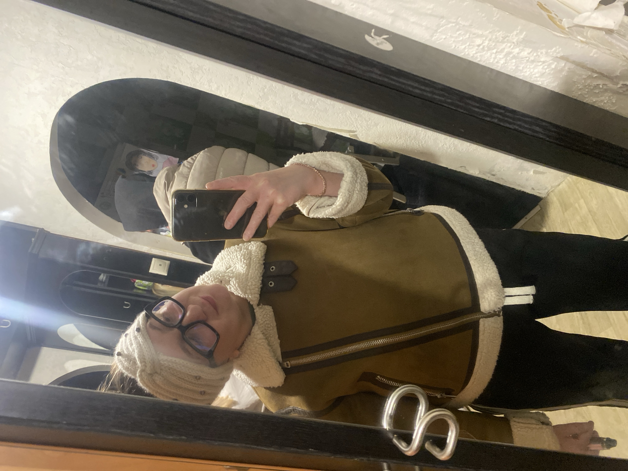 An indoor, mirror selfie photograph taken in Chaikovsky, Russia, features a female individual capturing her reflection. The subject is smiling faintly, wearing a light beige knitted beanie with dark embellishments and dark-framed eyeglasses. She is dressed in a brown aviator-style jacket with prominent white sherpa lining on the collar, cuffs, and hem, and a diagonal silver zipper. Her right hand, adorned with a delicate light-colored chain bracelet, holds a dark-colored smartphone, presumably an iPhone, up to take the picture. She is also wearing dark-colored pants with a visible white drawstring. The reflection reveals a textured, light-colored wall, an arched doorway, and a dark interior beyond, where a small picture or drawing is faintly visible. A light fixture is reflected above, casting a bright glare. The mirror itself has a dark, possibly black, frame. A light-colored floor is visible at the bottom of the frame.