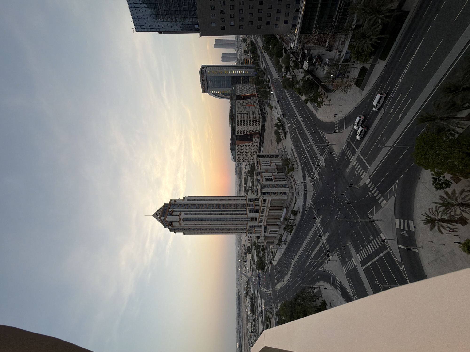 This is a high-angle, wide-shot photograph taken at dusk in Lusail, Qatar. The scene is dominated by a modern, multi-lane intersection with several vehicles visible, including cars and vans. Pedestrian crosswalks are clearly marked. In the background, a tall, ornate skyscraper with a distinctive pointed roof stands prominently, flanked by other contemporary buildings, some featuring glass facades. The sky exhibits the soft hues of sunset, with scattered clouds. Palm trees and other vegetation line the sidewalks and parts of the intersection, indicating a warm climate. The overall impression is one of an orderly urban environment during the transitional light of evening.