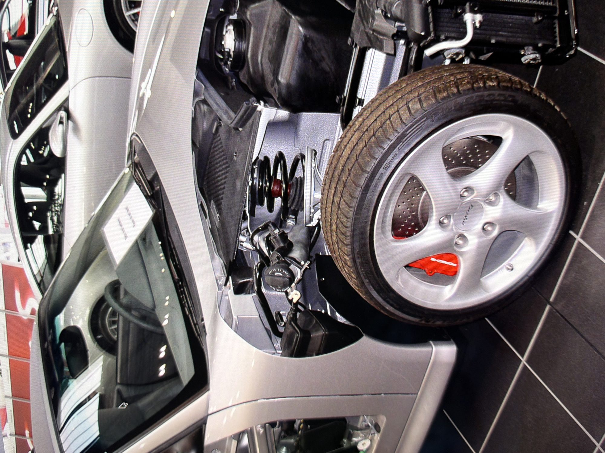 The image shows a close-up view of a silver Porsche sports car, partially disassembled, on display indoors. The car's front end is visible, with the hood open, revealing mechanical components like the suspension, engine parts, and a coil spring. A wheel with a silver rim and red brake calipers is prominently featured in the foreground. The car is situated on a dark tiled floor with white grout lines. In the background, other cars, including another silver Porsche and a red car, are visible, suggesting a car dealership or exhibition setting. There is no visible indication of the city of Kyiv, Ukraine, or any specific location within it.