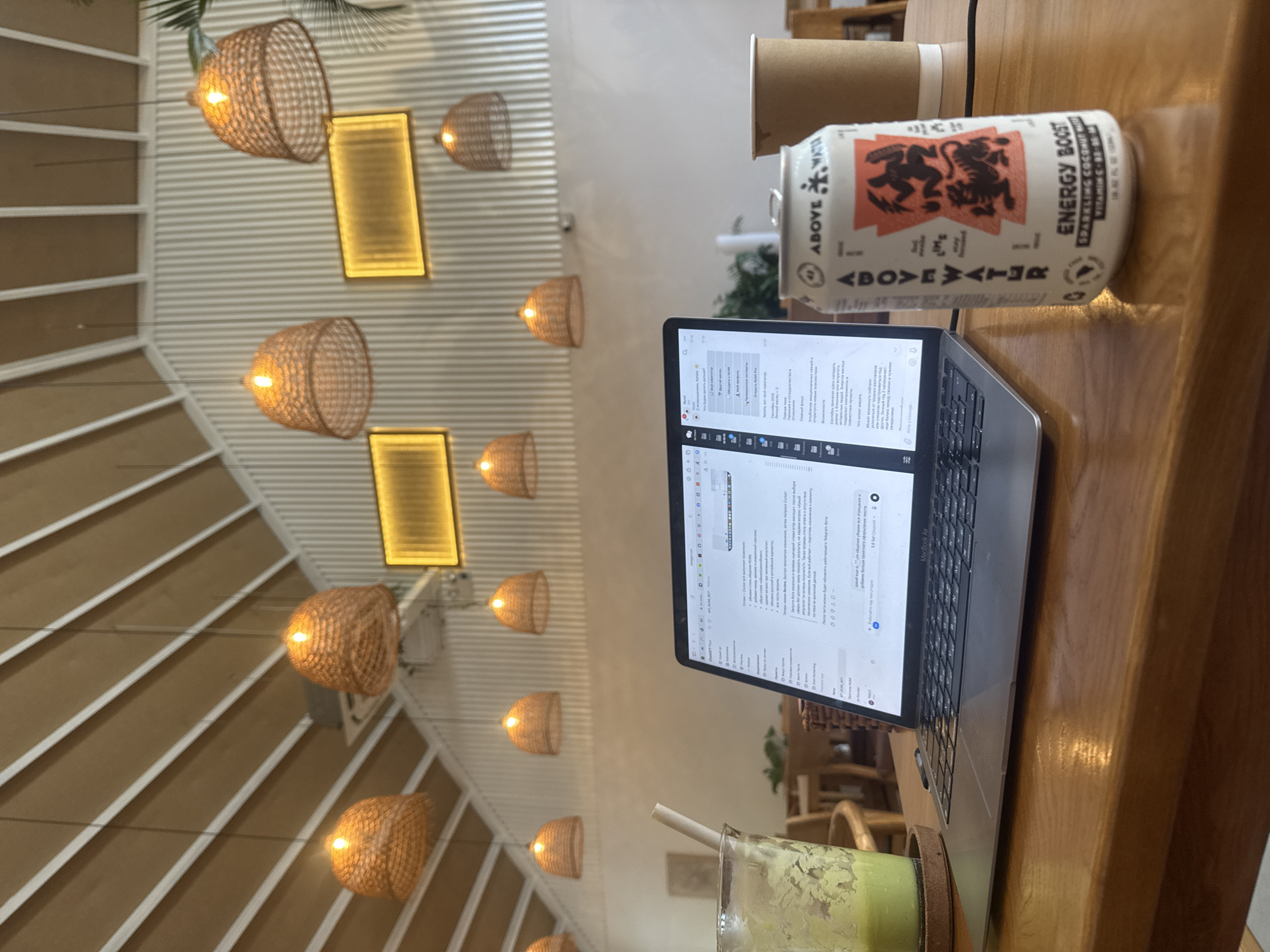 The image depicts a cozy indoor setting, likely a cafe or co-working space, with a focus on a work-from-home setup. The foreground is dominated by a wooden table on which a laptop, a can of "ABOVE WATER ENERGY BOOST" sparkling coconut water, and a cup of green liquid in a glass with a straw are placed. The laptop screen displays text in a foreign language, suggesting work or study is in progress. The background reveals an arched ceiling with exposed beams and several woven pendant lamps, casting a warm glow. Along the wall, there are two rectangular illuminated panels. The overall atmosphere suggests a productive and relaxed environment. While the image does not explicitly show the city of Thủ Đức, Vietnam, the style of architecture and decor, particularly the woven lamps and the ambient lighting, are commonly found in cafes and contemporary establishments throughout Vietnam.