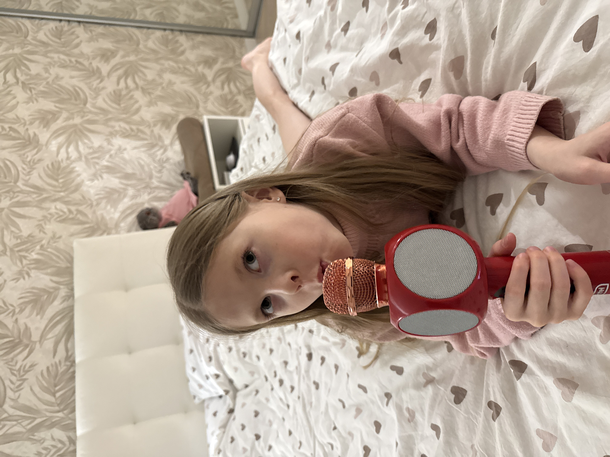 A young, light-skinned child with blonde hair lies on a white bedspread patterned with grey hearts. She is wearing a pink sweater. She is holding a red karaoke microphone close to her mouth. The headboard of the bed is white and tufted. The wallpaper behind the headboard is a beige and white floral print. A small shelf with items is visible to the left of the headboard. It is not possible to verify the location of the image or if it is Izhevsk, Russia.
