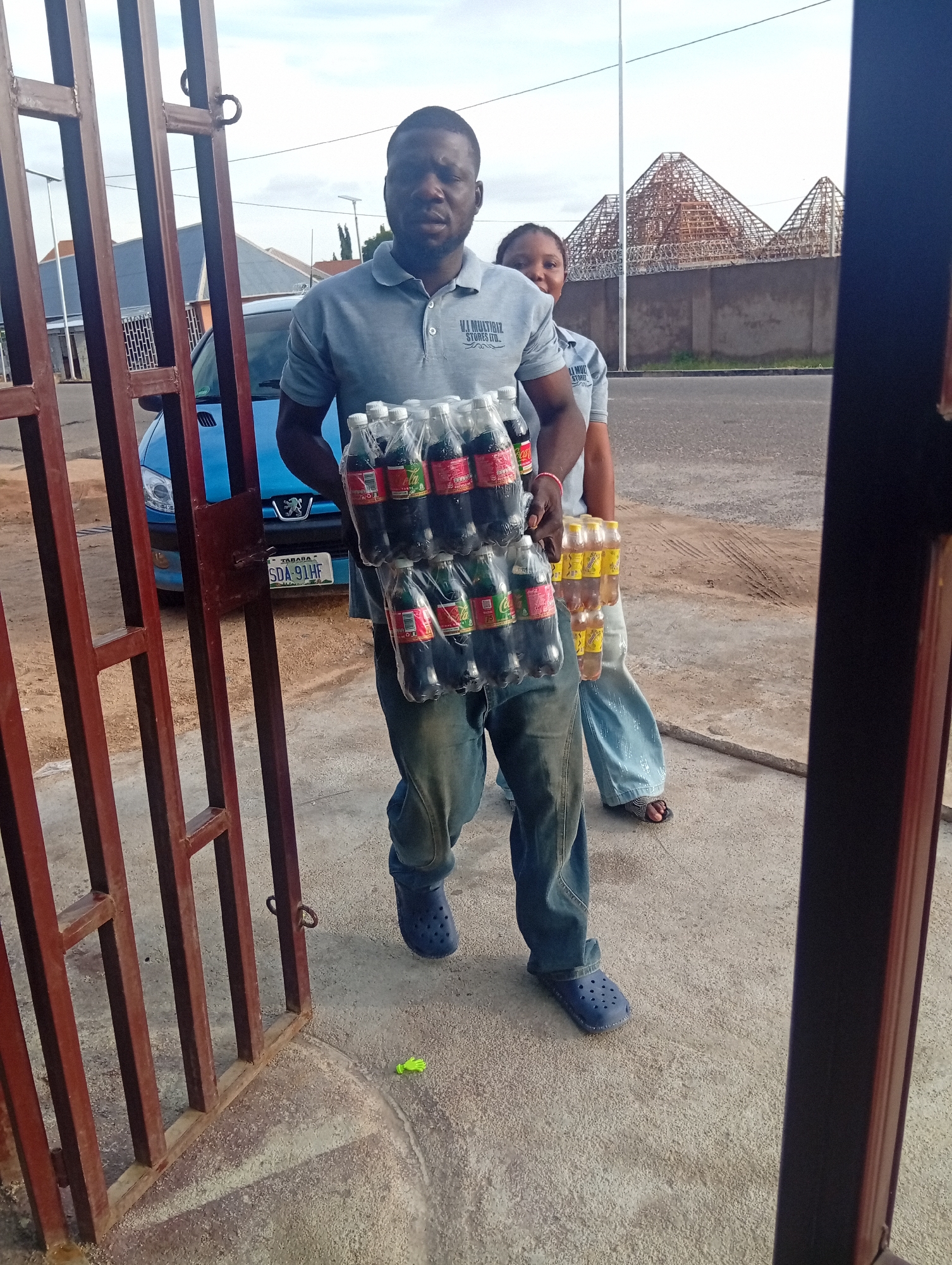 A man in a grey polo shirt and blue jeans carries a rack of soda bottles. He is walking through a metal gate. Behind him, a woman in light blue jeans and a grey t-shirt is partially visible. A blue Peugeot car is parked behind the man. In the background, a building with a distinctive pyramid-like roof structure is under construction. The scene appears to be in an outdoor urban setting. A green object, possibly a glove, lies on the ground near the gate. The license plate on the car reads "TABARA SDA 911HF". The sky is overcast.