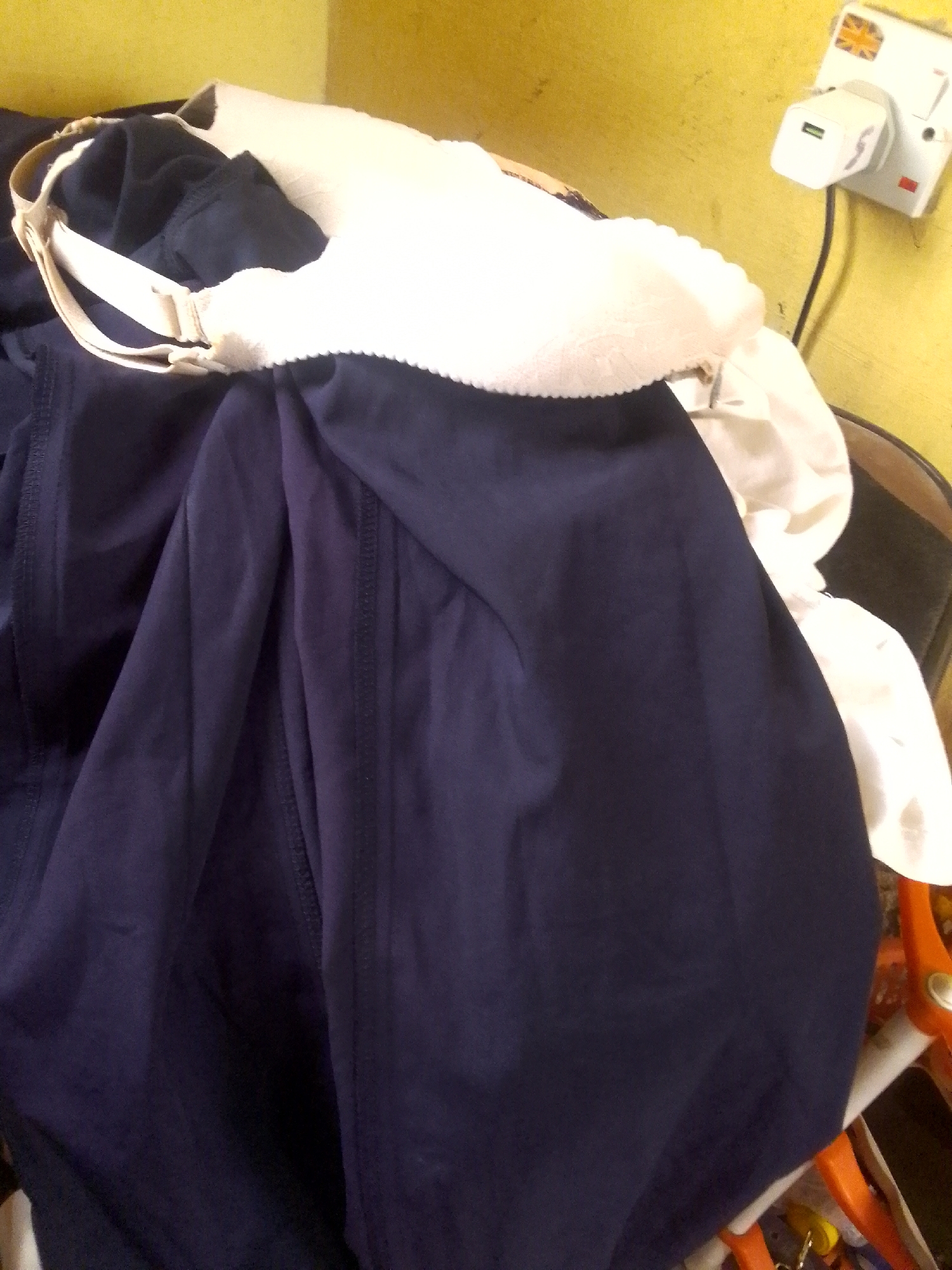 The image shows a close-up view of a dark blue garment, possibly a dress or skirt, draped over a white object, which appears to be a bra. The garment has prominent vertical seams running down its length. The bra has a beige strap and a ruffled edge. The background is a yellow wall with a white electrical outlet and a black cord. A small sticker with the Union Jack flag is visible on the outlet. The overall lighting suggests an indoor setting. There is no discernible activity or event taking place, and no people are visible. The image is taken from a slightly high angle, looking down at the clothing. The scene is likely in a residential or domestic setting in Jalingo, Nigeria.