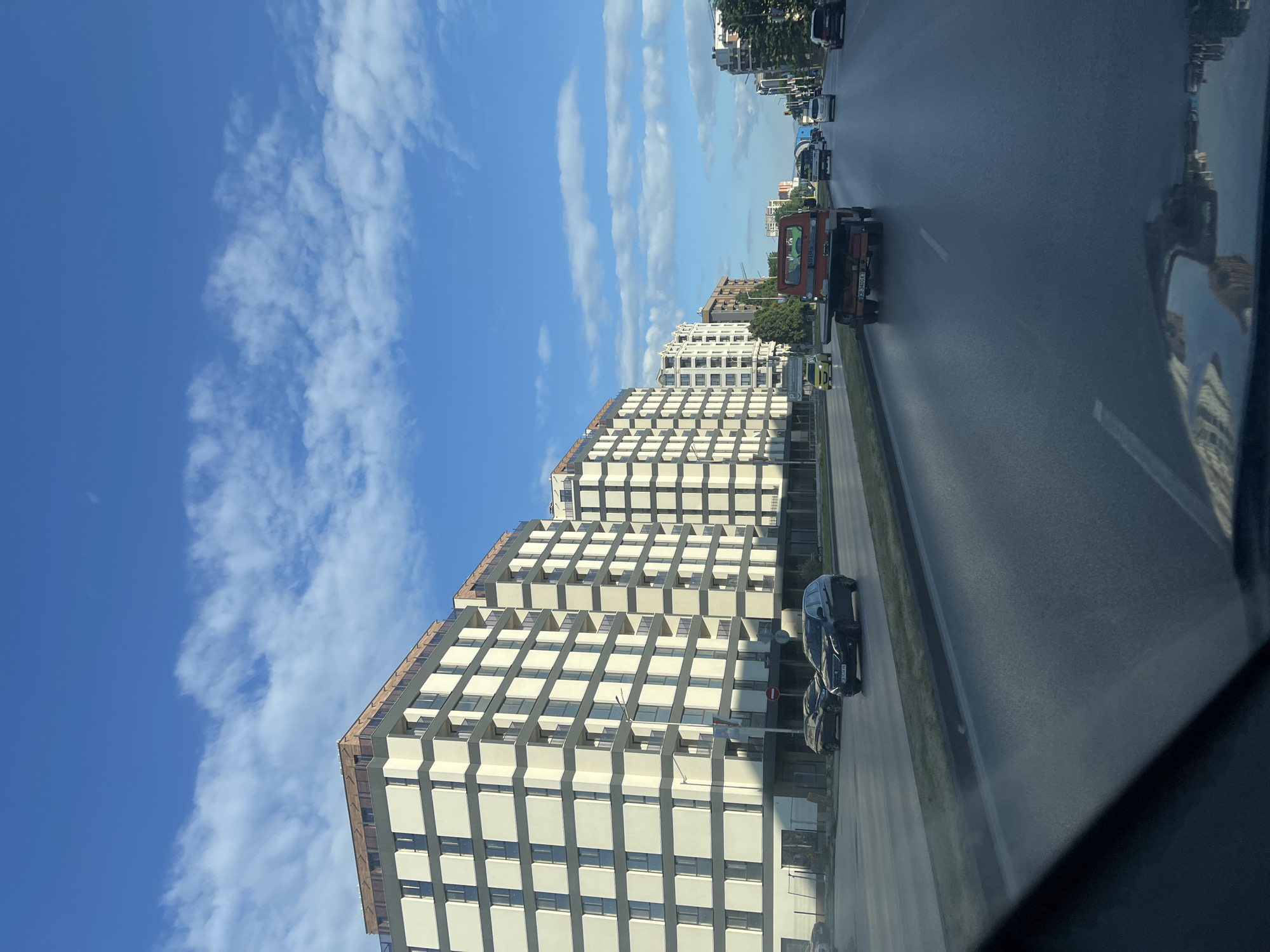 The image captures a street scene in a city under a partly cloudy blue sky. On the left side of the road, a row of modern, multi-story apartment buildings with white facades and dark grey accents lines the street. These buildings feature numerous windows and balconies. The road in the foreground is paved and appears to be a multi-lane thoroughfare with traffic. Several cars are visible on the road, including a black station wagon in the middle lane, a dark sedan to its left, and other vehicles further down the street. An orange tow truck with a flatbed is in the right lane, followed by other cars. Trees and traffic lights are visible along the sides of the street. The perspective suggests the photo was taken from inside a vehicle moving along the road.