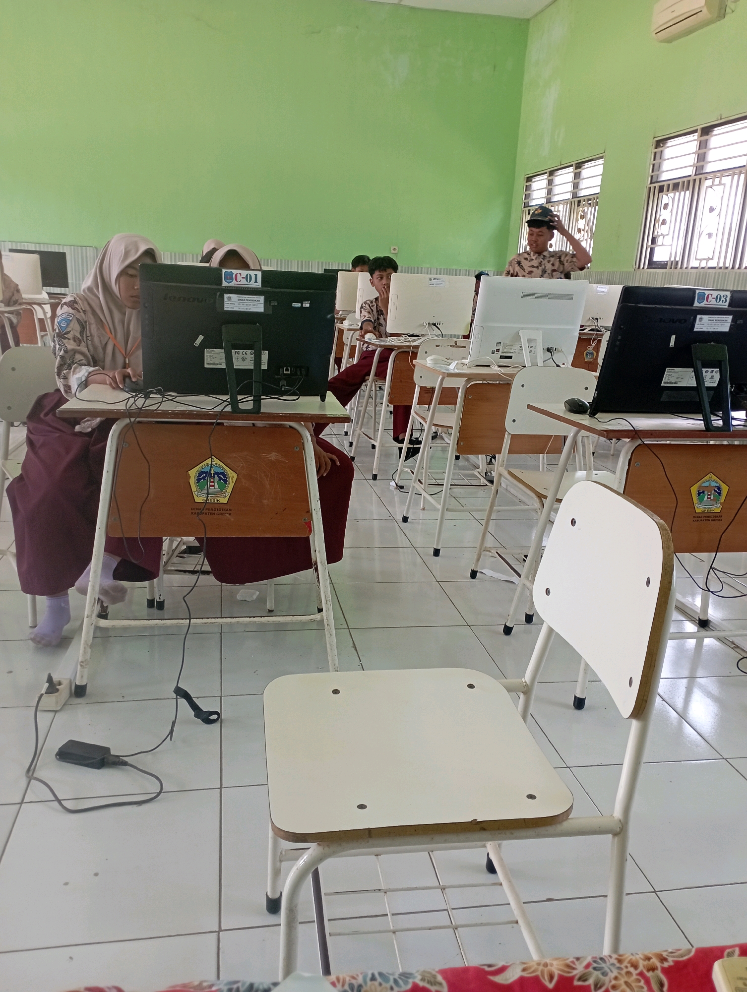 In a brightly lit classroom with pale green walls and tiled floors, several students are seated at individual computer desks. The desks have wooden panels with official emblems. Most students are focused on their computer screens, with some wearing headscarves and school uniforms. In the background, a male student in a camouflage uniform stands, adjusting his hat. The room is equipped with multiple desktop computers, chairs, and electrical cords. The scene appears to be a computer lab at a school in Banyuurip, Indonesia, likely during a lesson or examination.