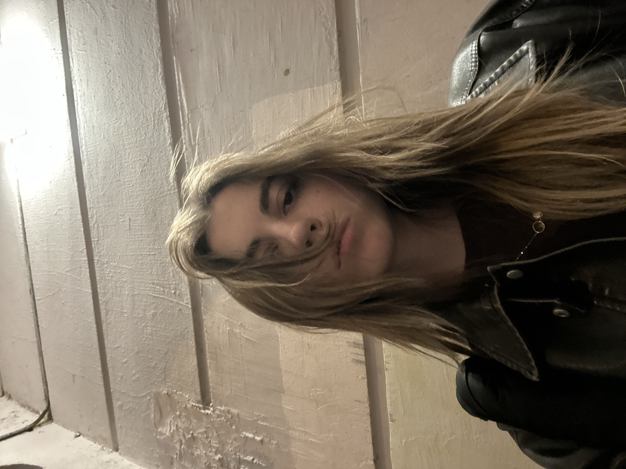 A young woman with long, brown hair, partially covering her face, is looking directly at the camera. She is wearing a dark brown top and a black leather jacket. A thin gold necklace with a small pendant is visible around her neck. She is positioned in front of a wall with horizontal planks, painted a light beige color. The lighting is dim, casting shadows and highlighting the texture of the wall. The overall impression is a candid, possibly indoor shot. The location is not specified in the image itself, therefore it cannot be identified as Moscow, Russia.