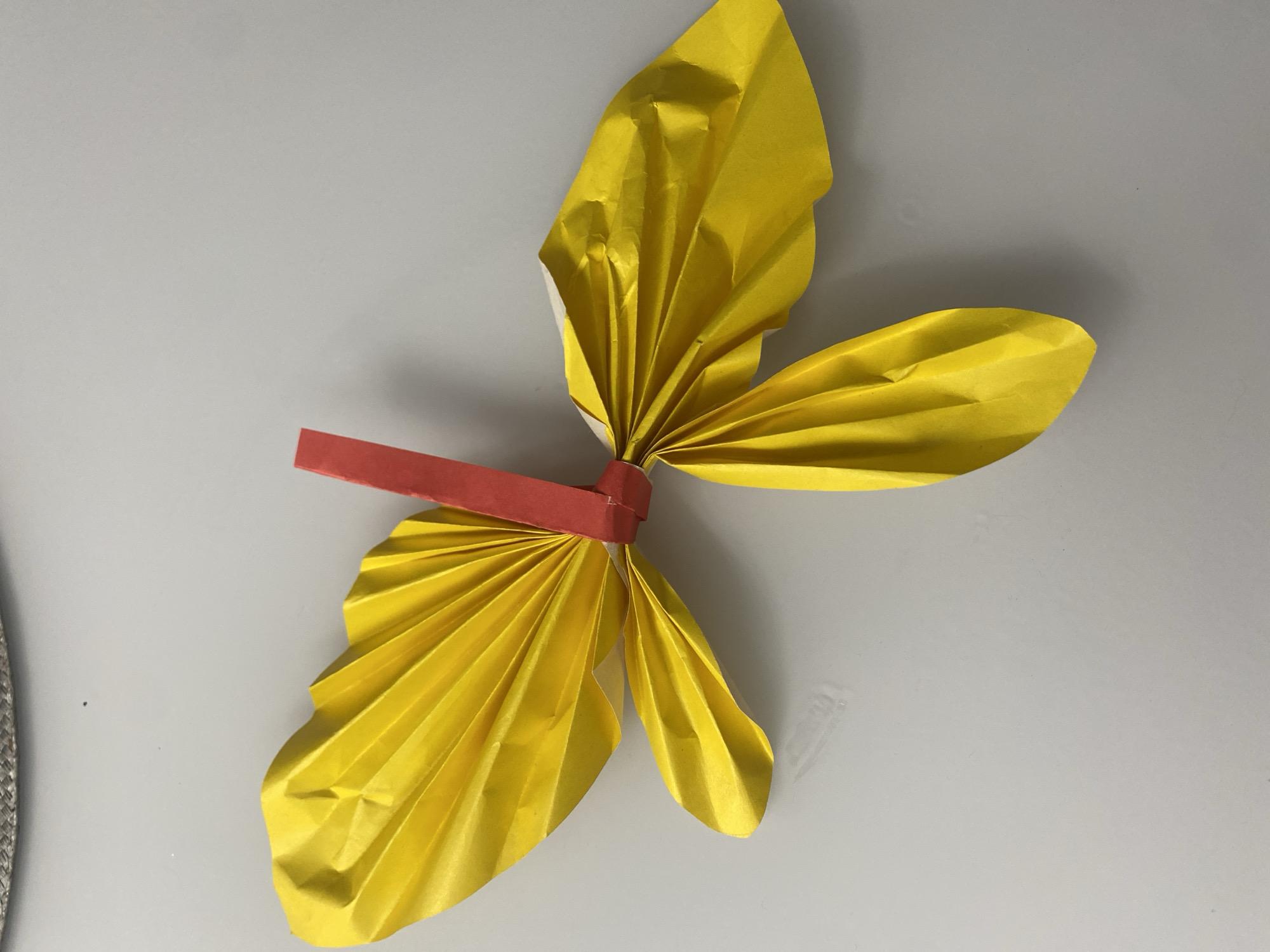 The image displays a handcrafted paper object resembling a butterfly or a stylized flower, positioned on a light gray or off-white surface. The main body of the object is formed from bright yellow paper, which has been accordion-folded to create a pleated texture, suggesting four distinct wings or petals. These yellow sections fan out from a central point.
Securing the yellow paper wings at their center is a slender strip of red paper, wrapped horizontally around the folded yellow sections, forming what appears to be the body of the butterfly or the center of the flower. This red strip has a slight sheen, possibly from the paper type or lighting.
The background is a plain, even-toned surface, likely a tabletop or wall, with subtle shadows cast by the paper craft, indicating a light source from above and to the left. No discernible activity is taking place; the image is a still-life shot of the completed craft. There are no people visible, and no text is present in the image. The scene appears to be an indoor setting, possibly in Izhevsk, Russia, where the craft might be displayed or photographed. The lighting is soft and consistent, highlighting the texture and colors of the paper.