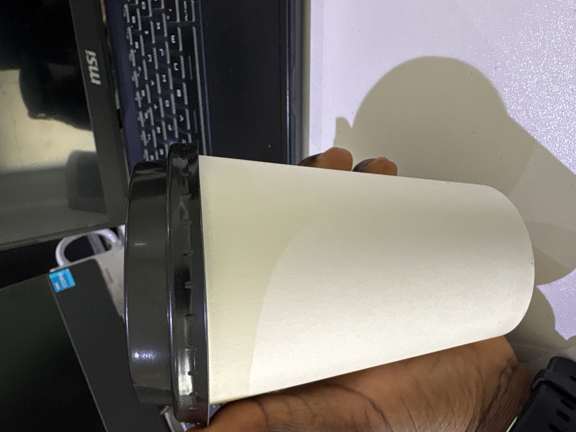 A close-up, slightly rotated shot captures a dark-skinned hand holding a plain white disposable cup with a black lid. The cup is held horizontally, positioned centrally in the frame. A black laptop, oriented vertically, is partially visible in the background, with its screen displaying the "MSI" logo prominently in white letters on its bezel. To the right of the laptop screen, part of the keyboard with visible keys is seen. Further to the left and slightly below the laptop, a device with a blue sticker, likely a brand emblem such as Intel, is visible. The hand holding the cup also wears a black smartwatch or fitness tracker on the wrist.
The setting appears to be indoors, likely at a desk or table, with the laptop and cup placed on a light-colored surface. A distinct, large shadow of the cup and hand is cast upon a light-colored wall or background surface to the right, suggesting the presence of a light source from the left. The overall lighting is indicative of an indoor environment. No specific activity beyond holding the cup is discernible, but it suggests the person might be about to drink. The scene is located in Gwaska, Nigeria.