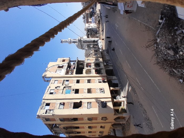 This image depicts a street scene in the city of Khan Yunis, Palestinian Territory, characterized by significant structural damage to buildings. The view is angled downwards, framed in the upper portion by a thick, rope-like object extending diagonally from left to right.
To the left, a multi-story, light-beige residential or commercial building stands extensively damaged, with multiple large holes penetrating its facade, exposing interior rooms and structural elements. Windows are broken or missing, and fabric or debris hangs from various openings and balconies. Debris piles are visible at the base of the building.
The asphalt street, with visible white lane markings, runs through the center of the frame and appears relatively clear. Several individuals, including some children, are visible walking on the street and sidewalks. A child is seen running on the left side of the street. In the middle distance, several people are gathered or seated on the sidewalk next to piles of rubble.
Further down the street, on the right side, a white mosque with a prominent minaret and dome is visible, surrounded by substantial rubble and damaged structures. The area around the mosque also shows widespread destruction. Piles of rubble, concrete fragments, and dry vegetation are present in the lower right foreground. The sky above is clear and blue.