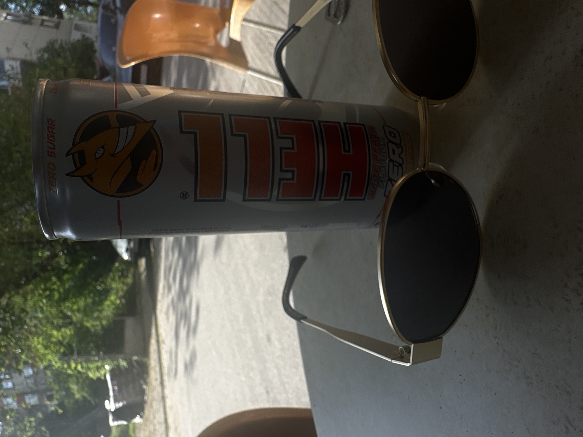 The image depicts a can of "HELL ZERO" energy drink placed on a table. On the right of the can are a pair of round, gold-rimmed sunglasses. The background shows a street scene with trees and buildings, possibly in an urban area. The photograph was taken outdoors, with natural light. Based on the buildings and environment, it is impossible to pinpoint the exact location in Plovdiv, Bulgaria, without additional information.