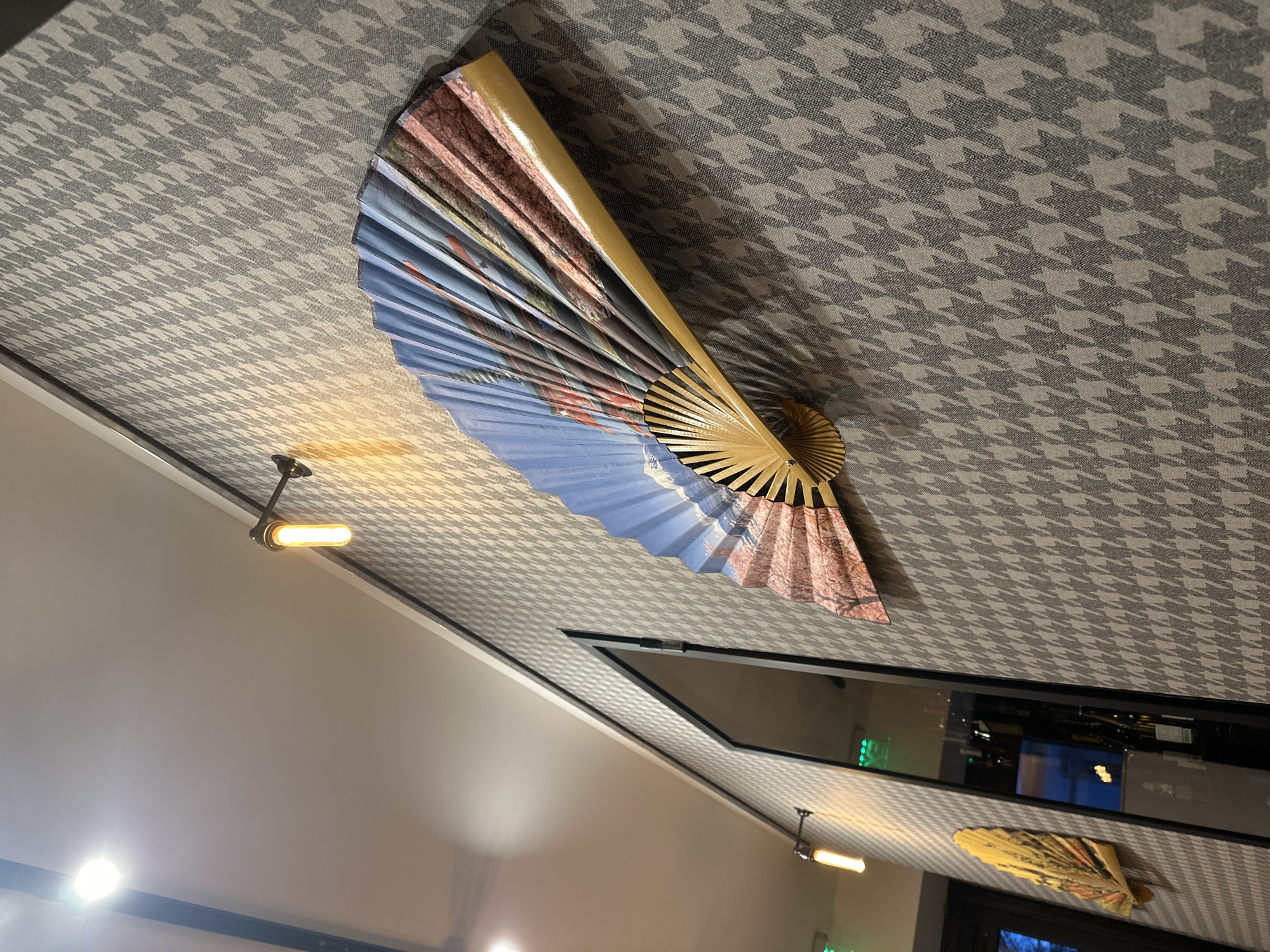 A medium shot captures a view of the ceiling of a building in Sofia, Bulgaria. The ceiling is covered with a grey and white houndstooth patterned material. On the ceiling is mounted an opened decorative fan, predominantly blue and pink. There is another similar decorative fan partially visible near a window in the lower right corner. A light fixture with a cylindrical bulb is also mounted on the ceiling near a white wall. The scene appears to be indoors.